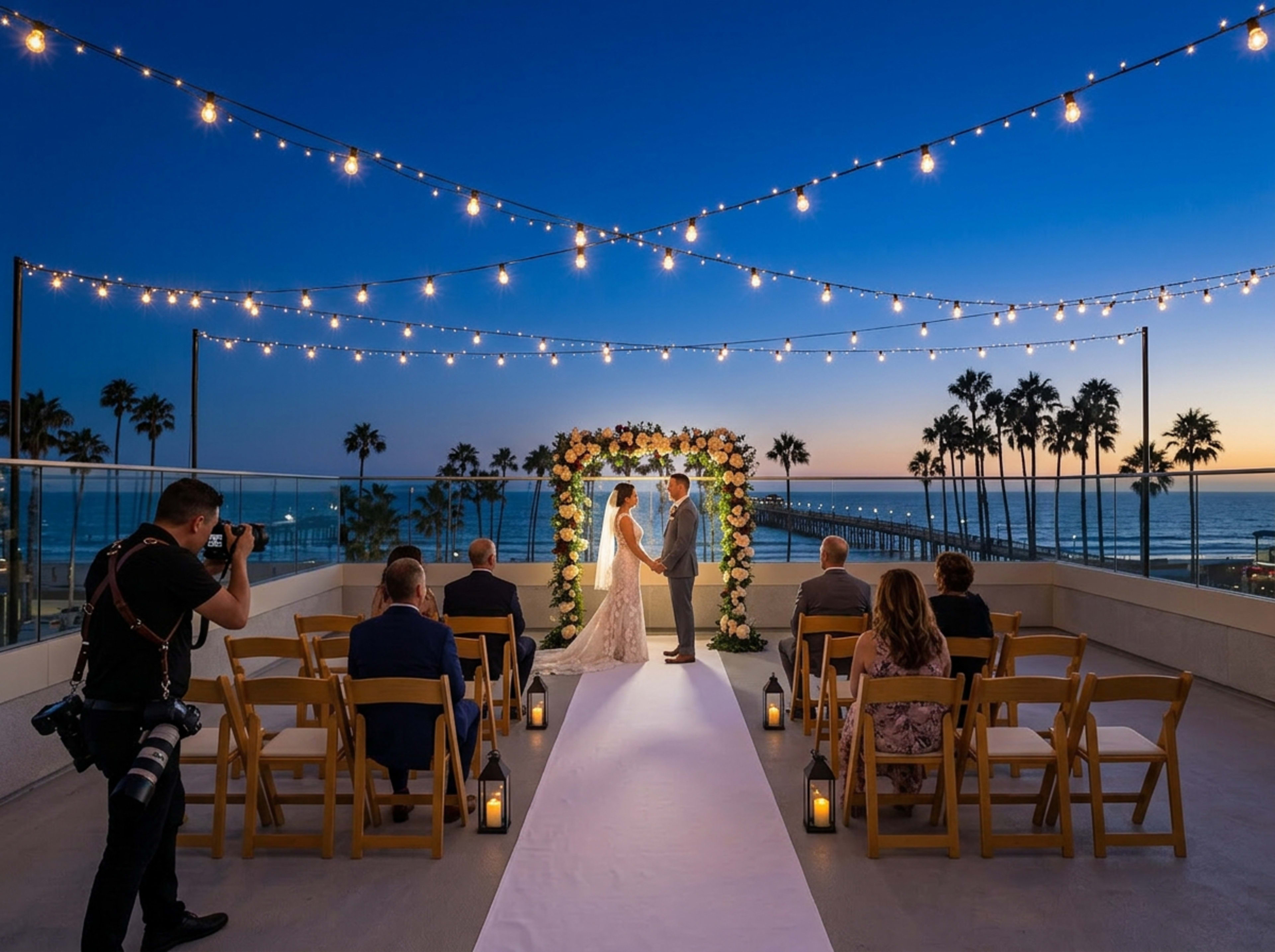 A couple stands under a floral arch exchanging vows during a sunset wedding ceremony on a rooftop, surrounded by guests seated in wooden chairs.