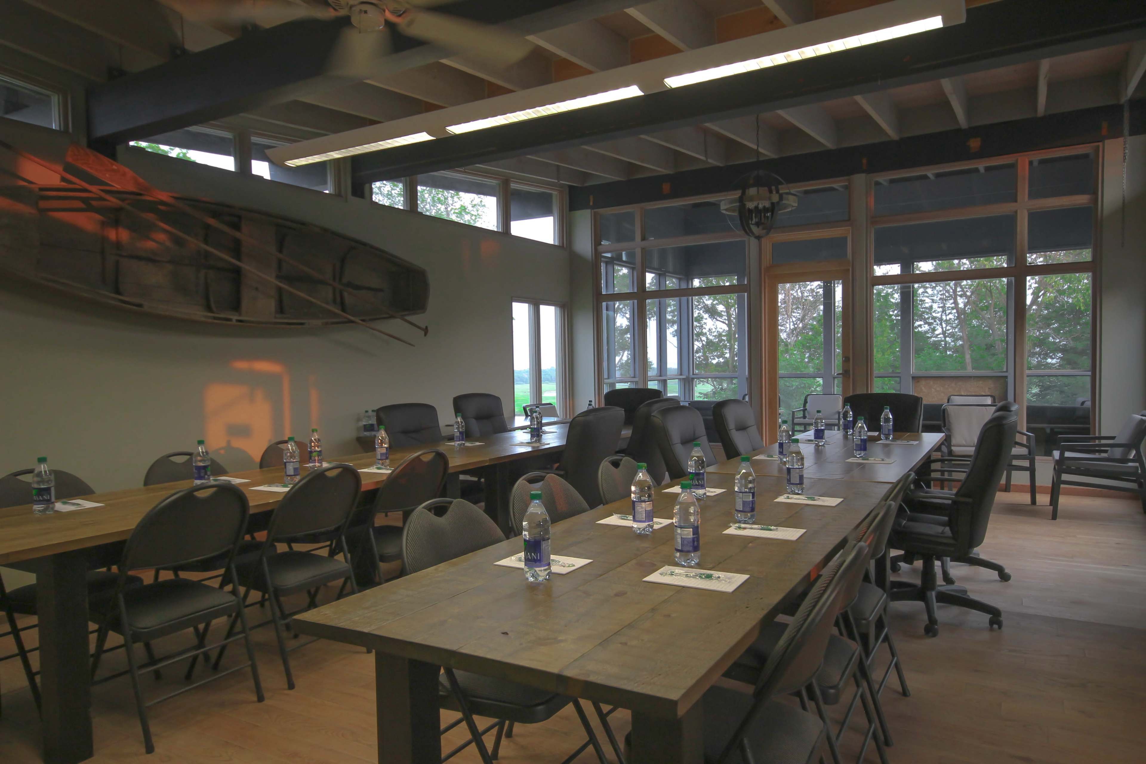 A meeting room features long wooden tables arranged with chairs, bottles of water, and large windows offering a view of the outdoors.