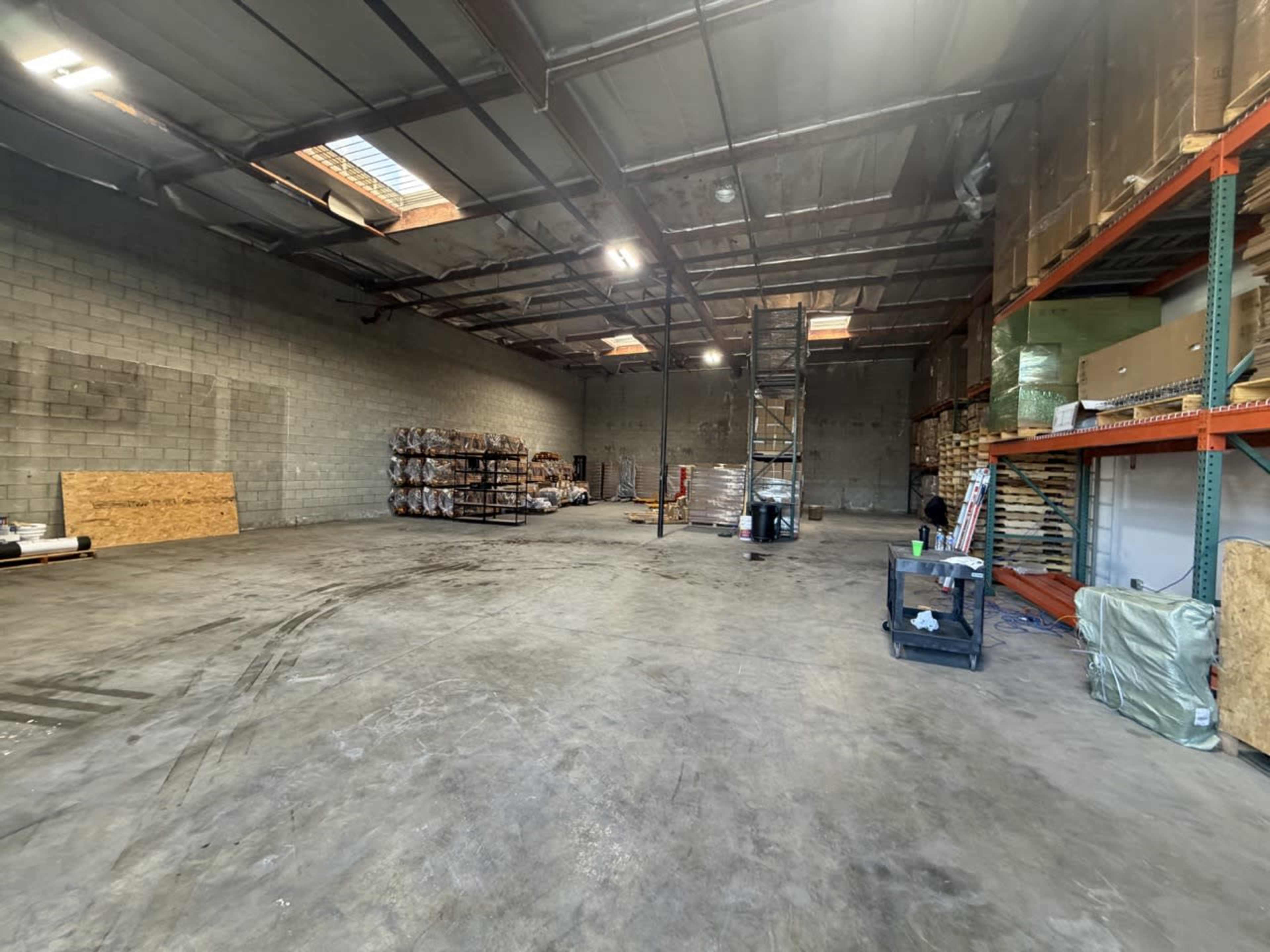 The image shows an empty warehouse interior with exposed concrete walls, tall ceilings, and shelves stocked with materials along one side.