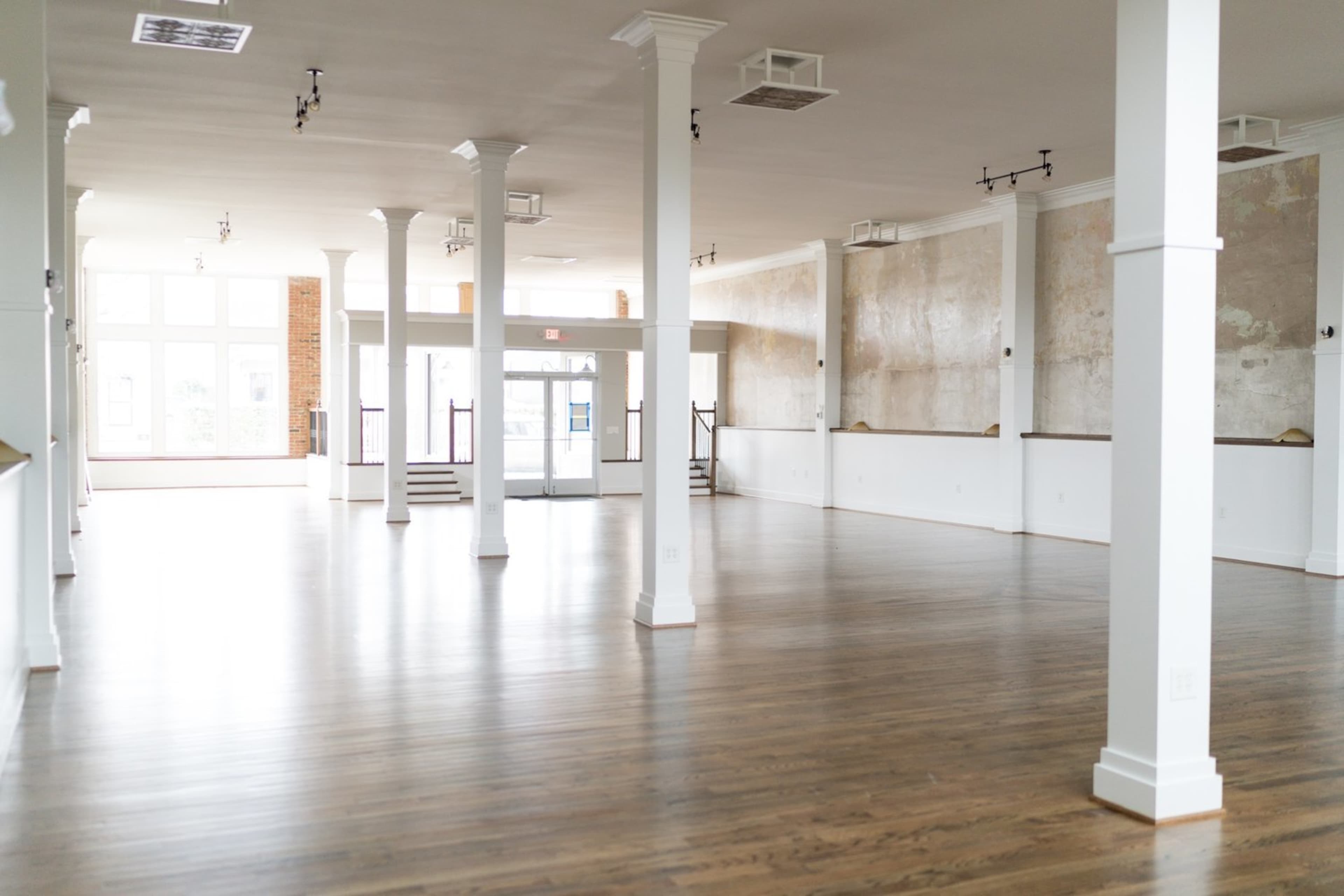 A spacious, empty hall with polished wooden floors, tall white pillars, and large windows letting in natural light.