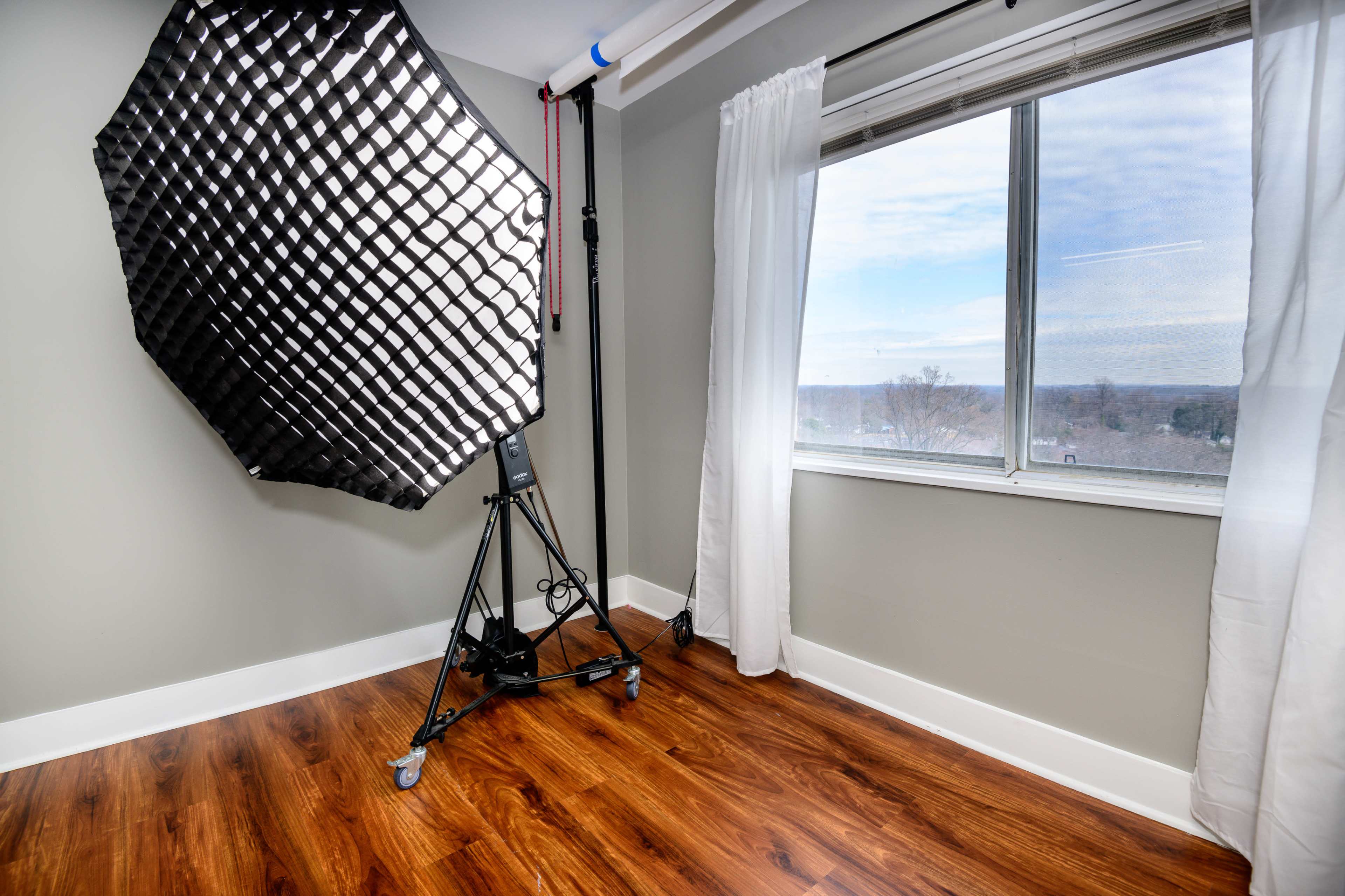 A brightly lit room with a large window, featuring a black softbox light on a stand and hardwood flooring.