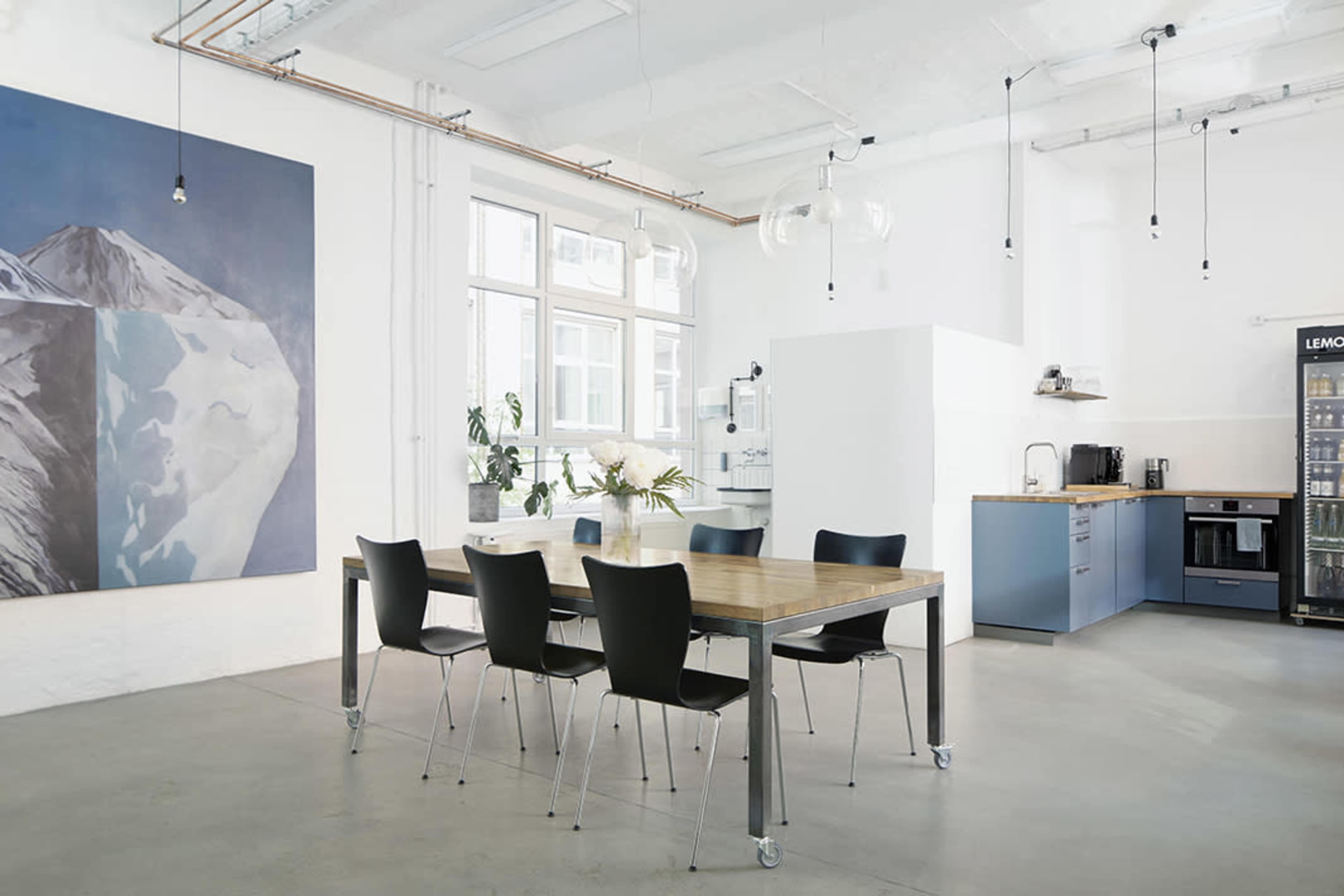 A modern kitchen and dining area features a wooden table surrounded by black chairs, with large windows and a mountain-themed artwork on the wall.