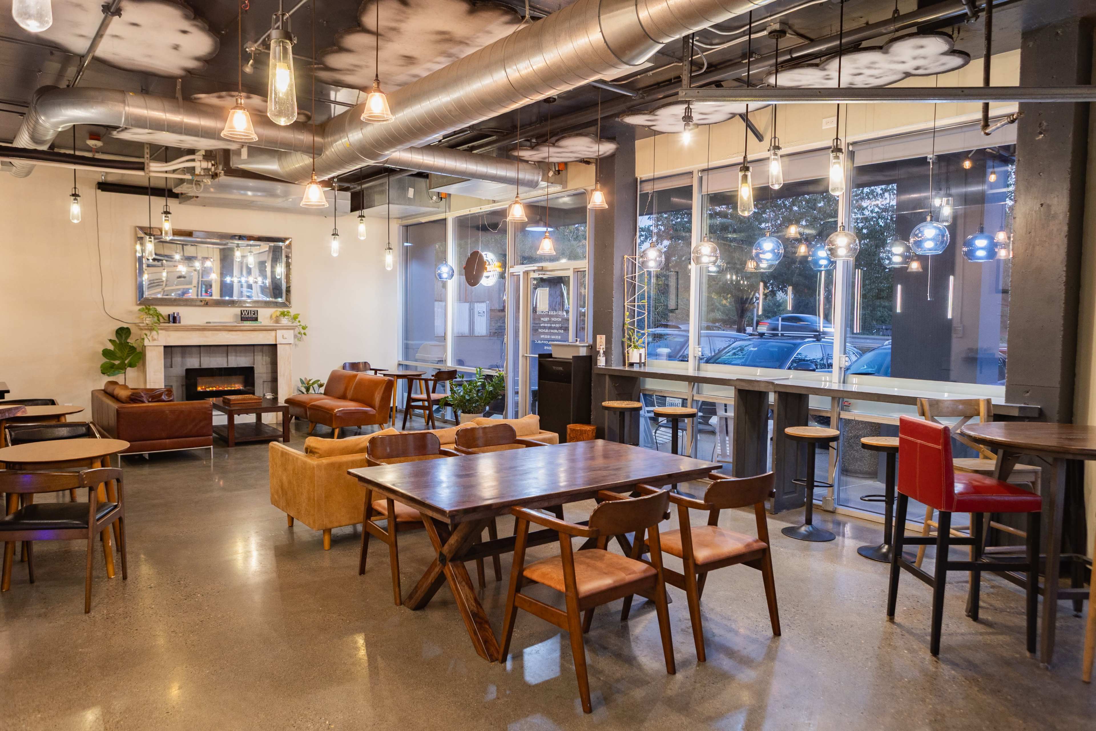 The image shows a modern cafe with wooden tables, varying seating arrangements, and large windows letting in natural light.