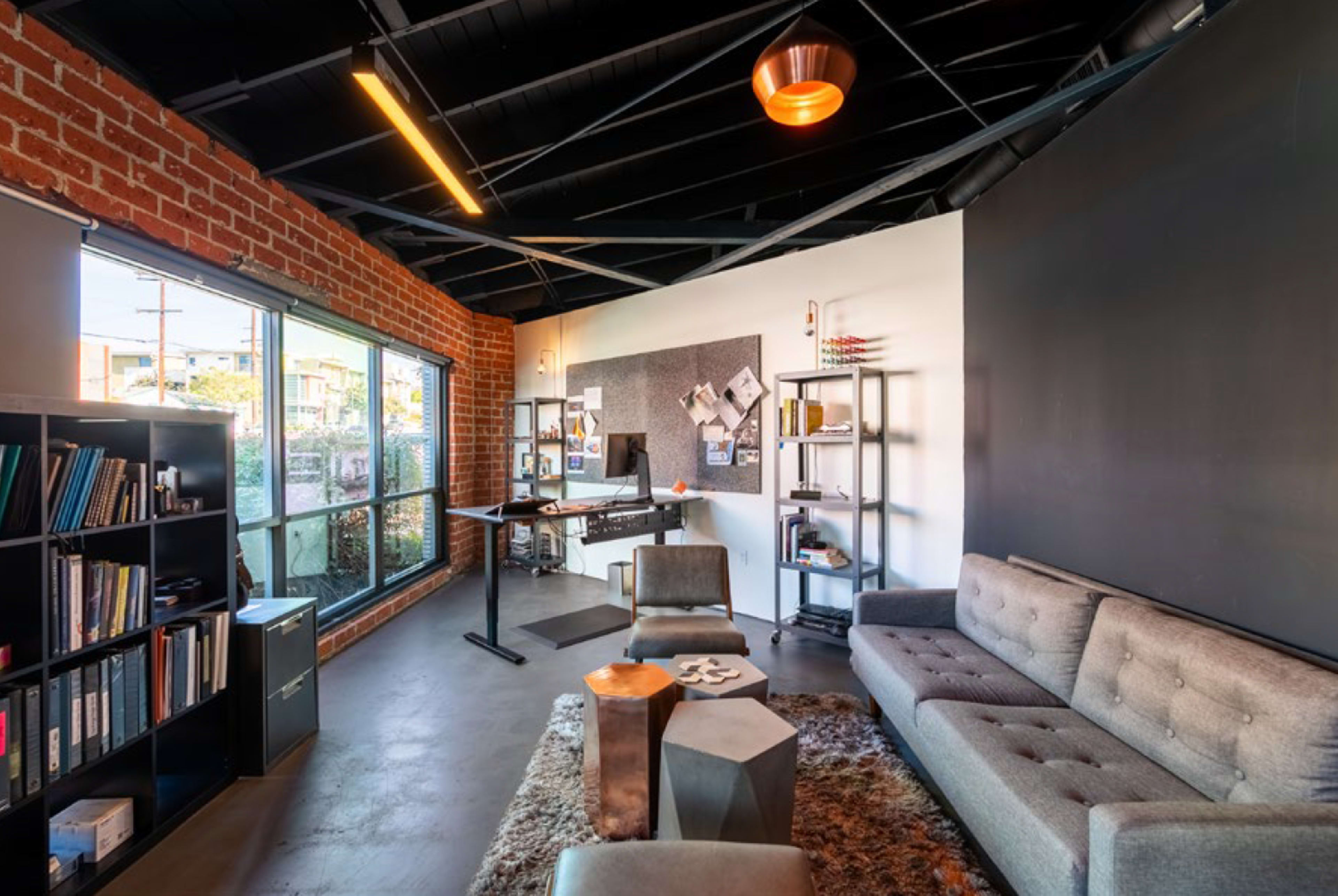 The image shows a modern office space featuring a gray sofa, a coffee table, shelves with books, and a desk with a view through large windows.