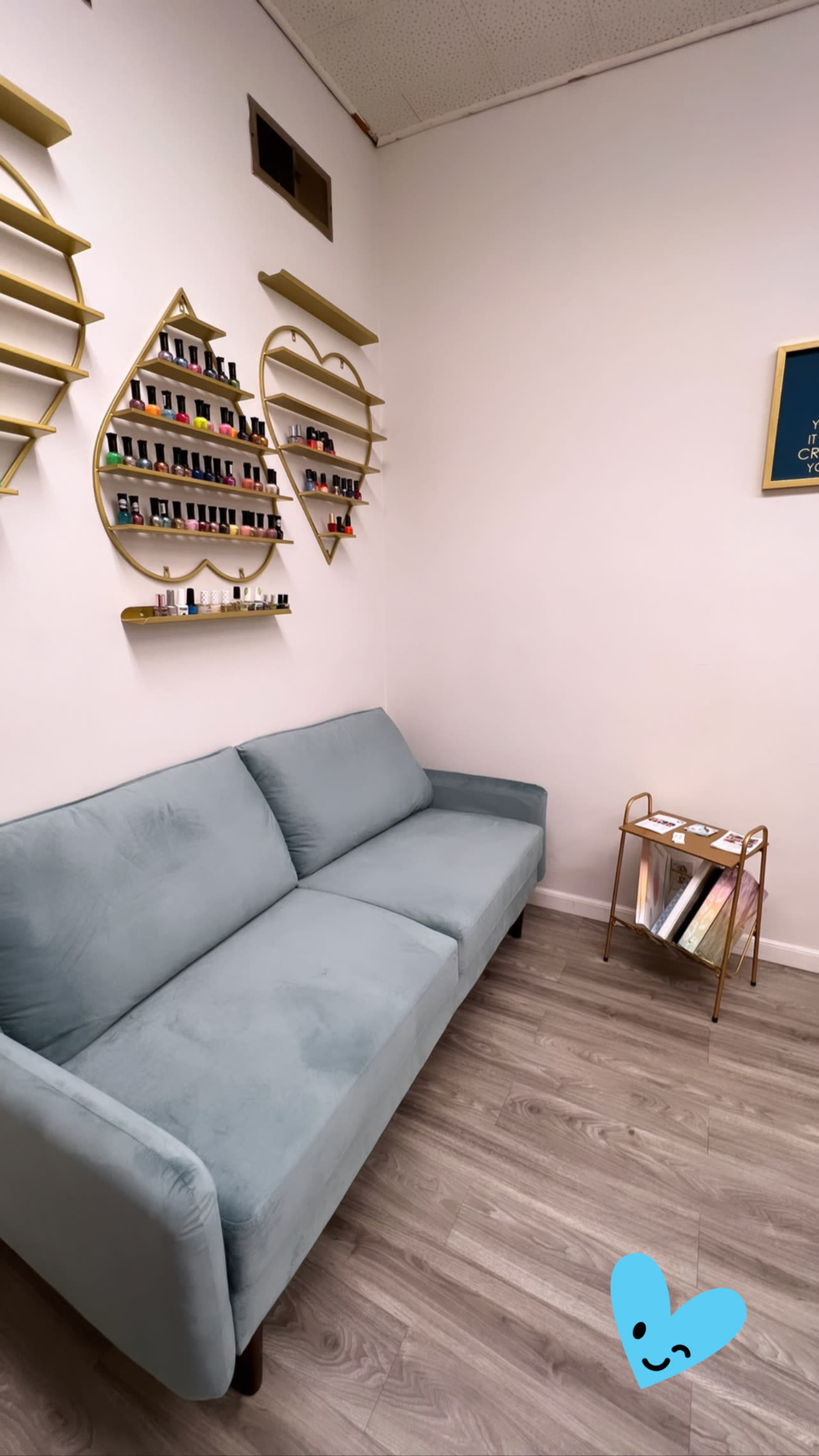 The image shows a small, well-decorated room with a light blue couch and shelves displaying various nail polish bottles on the walls.
