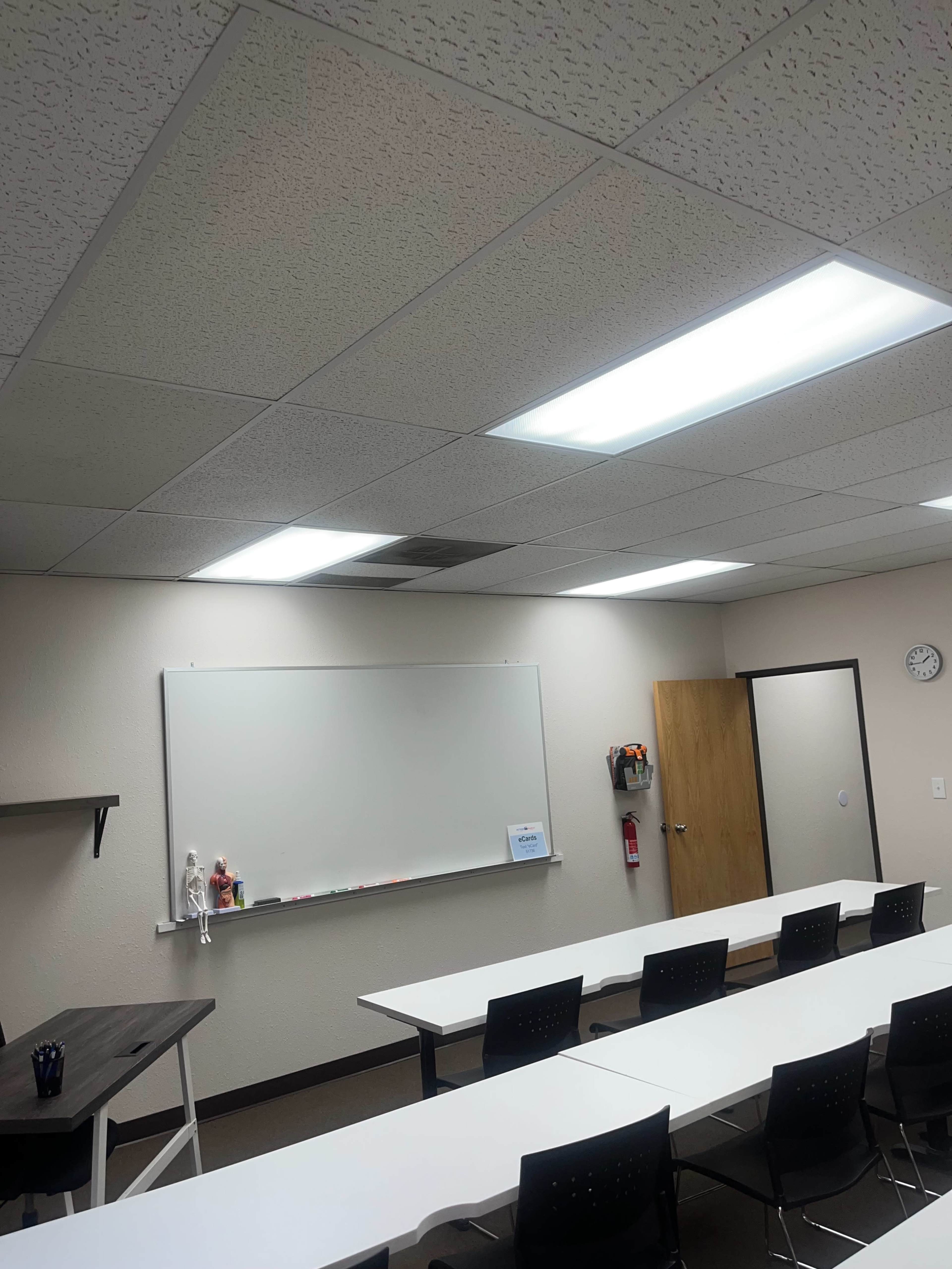 The image shows a classroom interior featuring a whiteboard, rows of tables and chairs, a clock on the wall, and overhead fluorescent lights.