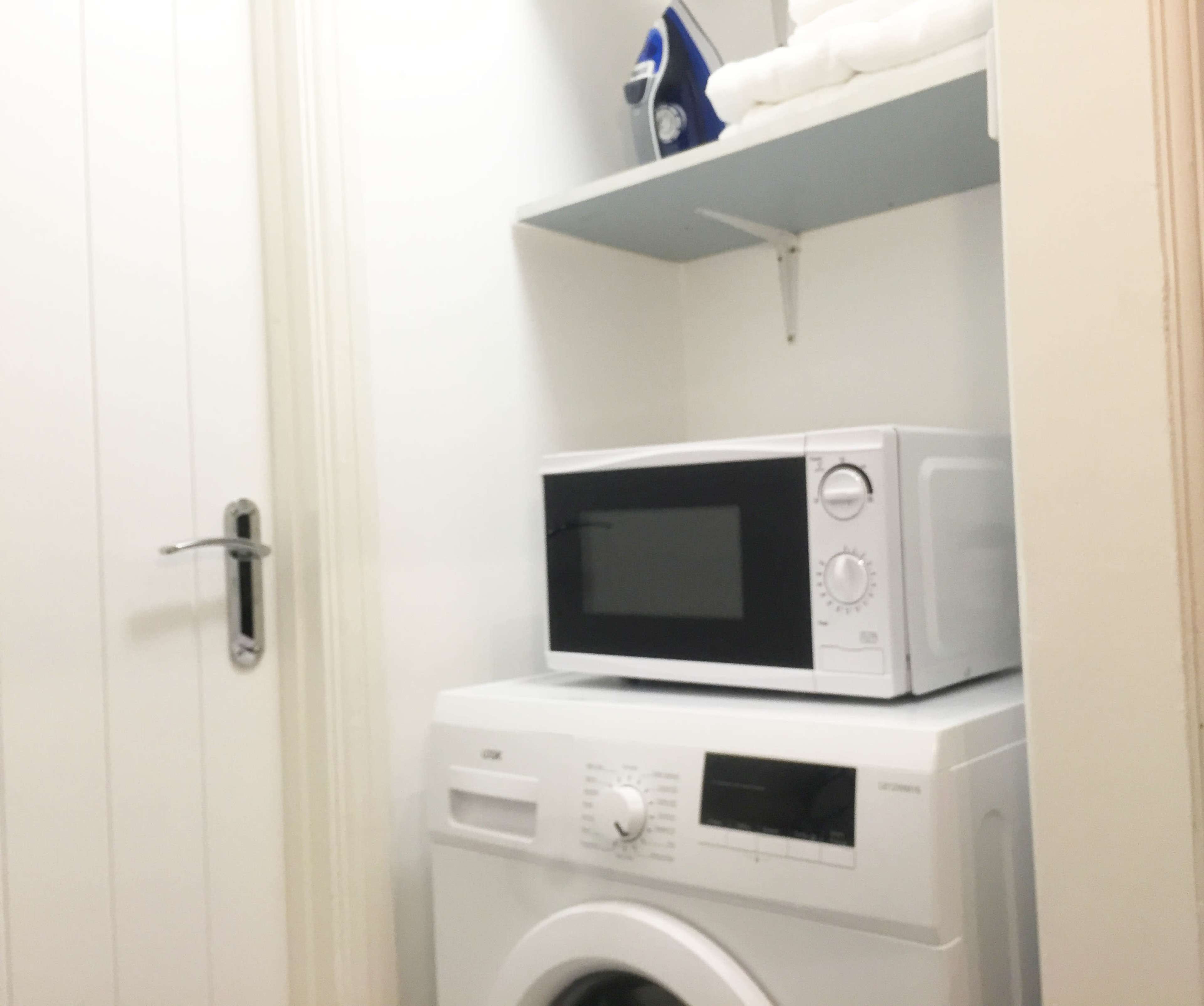 A laundry room with a washing machine beneath a microwave and an iron on a shelf.