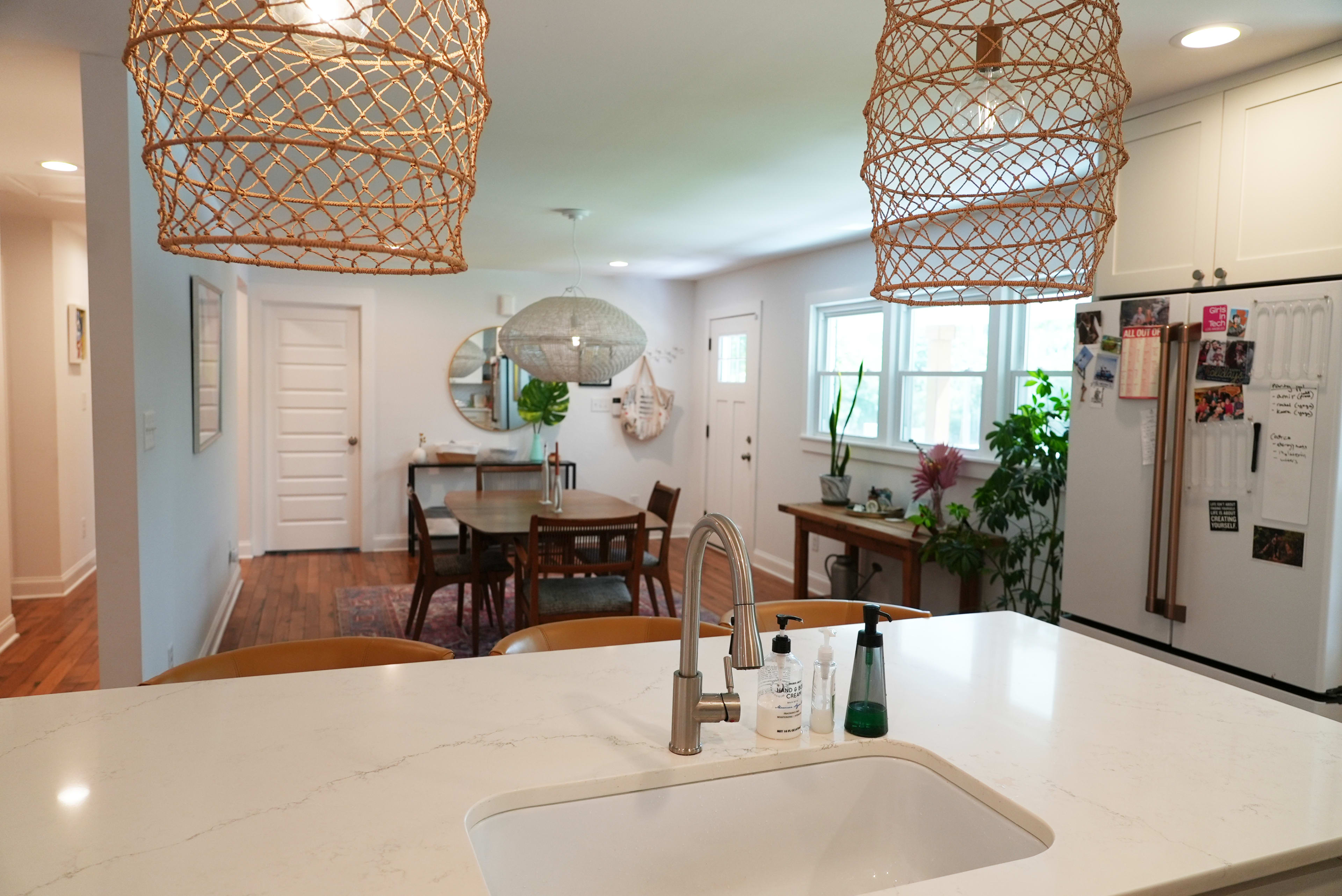 A modern kitchen with a white marble countertop, featuring two woven light fixtures, and an open dining area in the background with a wooden table and chairs.