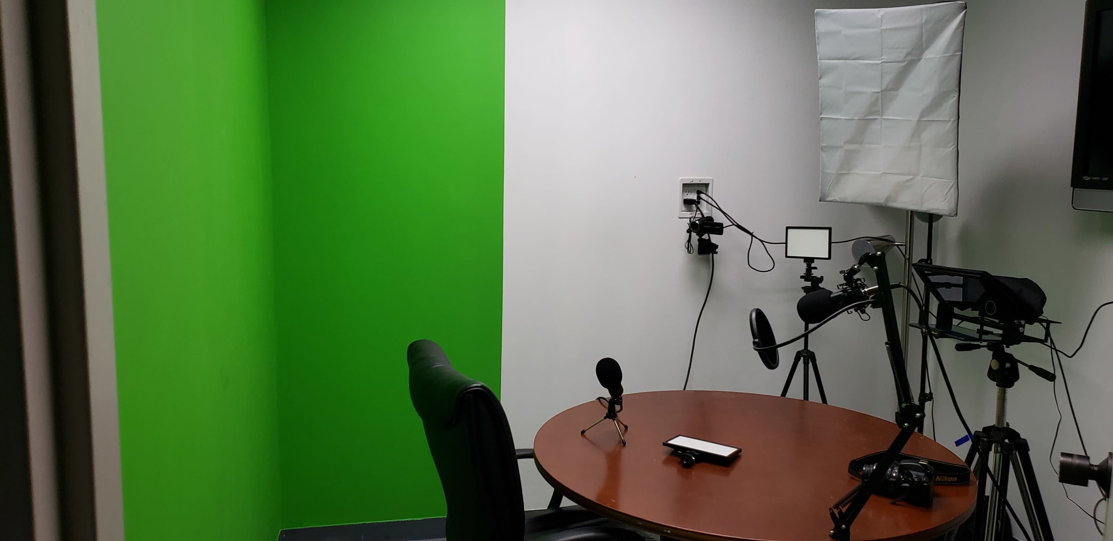 A recording studio setup with a green screen on one wall, a round table, and cameras positioned on tripods.