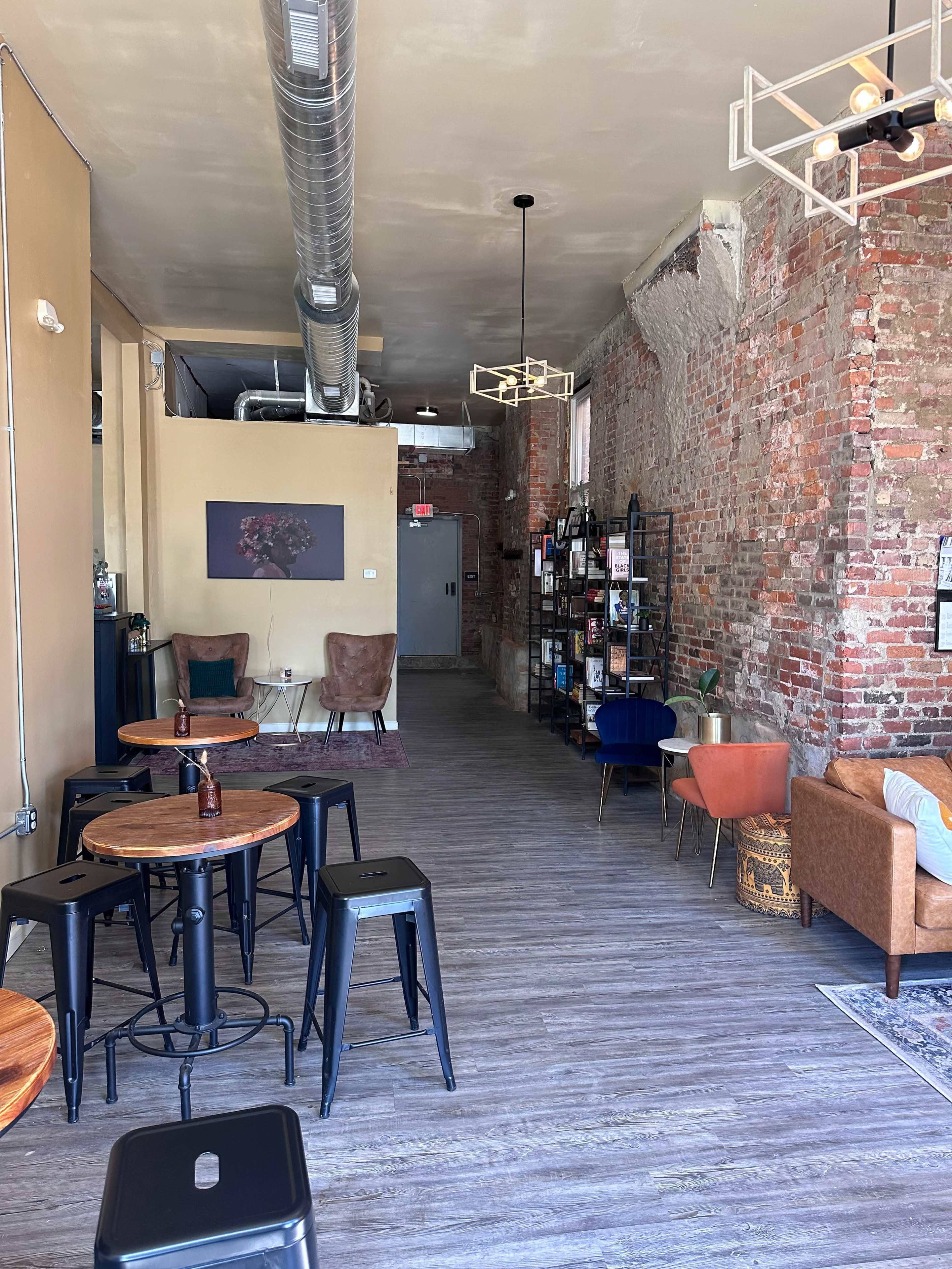 A modern café interior with exposed brick walls, wooden floors, and a mix of seating options including metal stools and upholstered chairs.