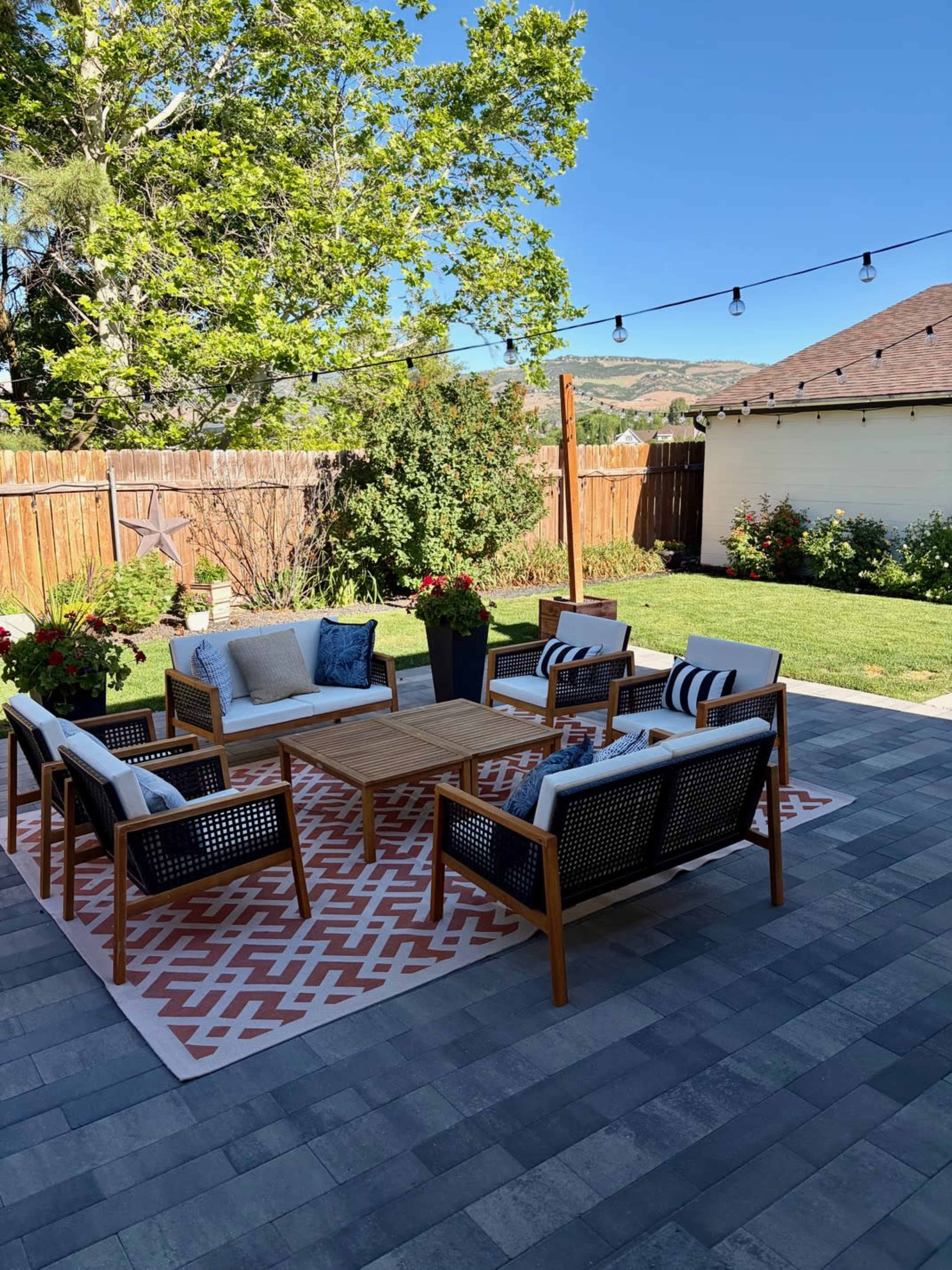 A furnished outdoor patio features a seating arrangement with four chairs and a central table, surrounded by greenery and string lights above.