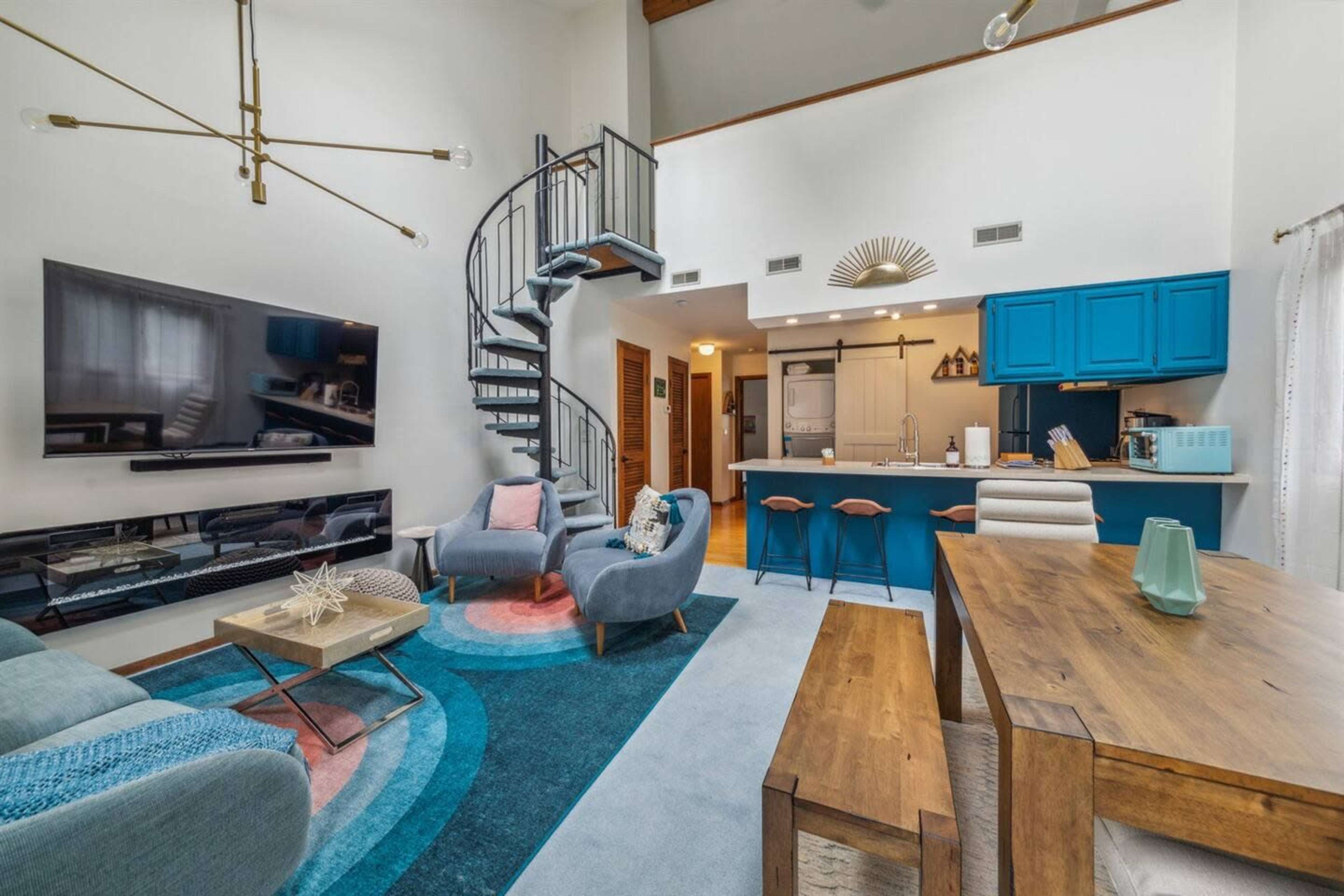 A modern living space featuring a spiral staircase, a blue kitchen, and a cozy seating area with a large television and a dining table.