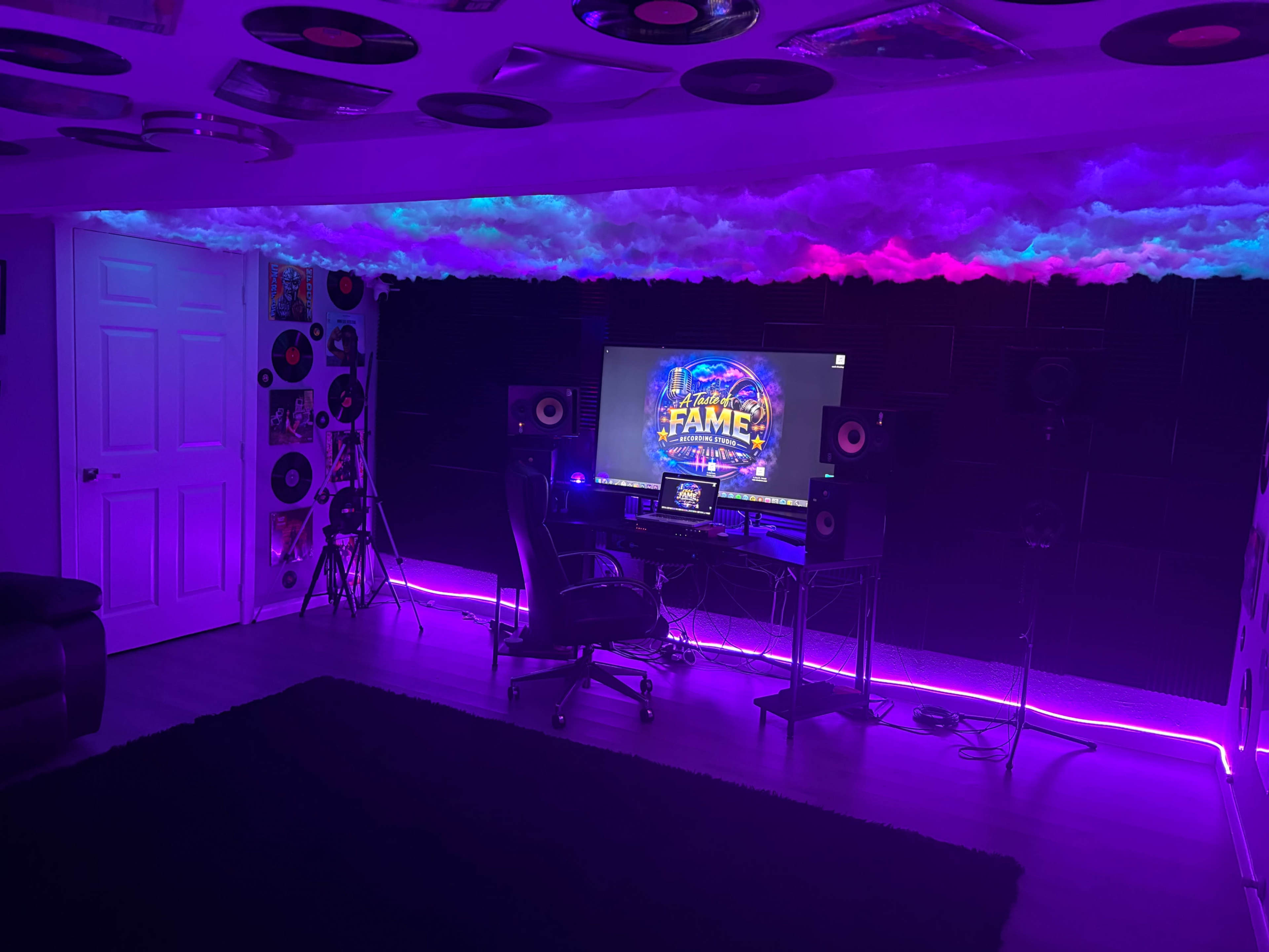 A home studio features a computer setup surrounded by colorful lighting, sound panels, and vinyl records on the walls.
