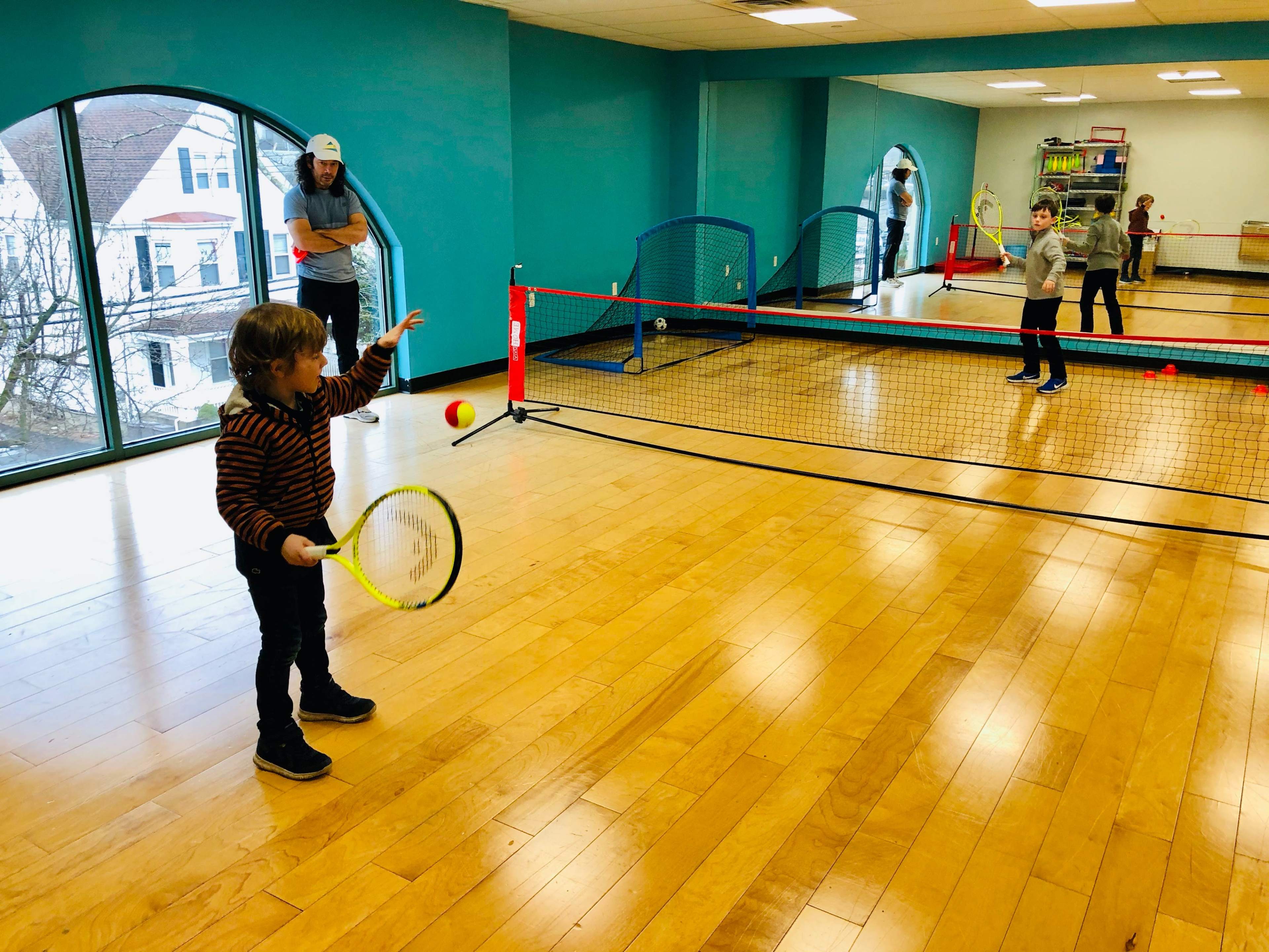 A young child prepares to hit a tennis ball while another child stands at the opposite side of a small indoor court.