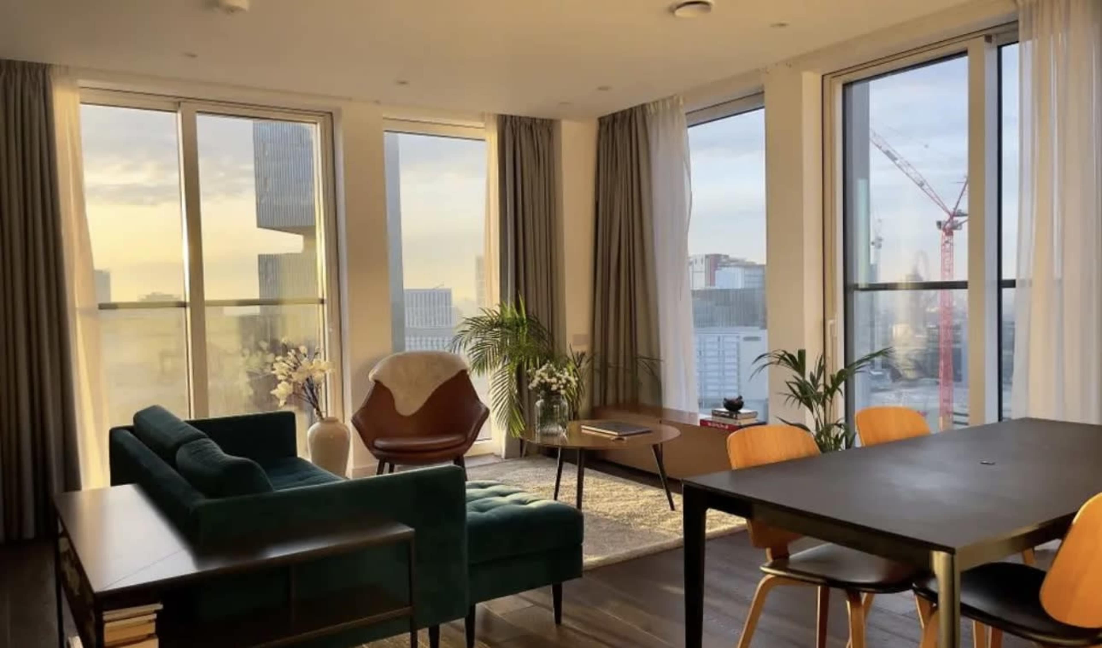 The image shows a modern living space with large windows, a green sofa, a wooden dining table, and a view of city buildings outside.