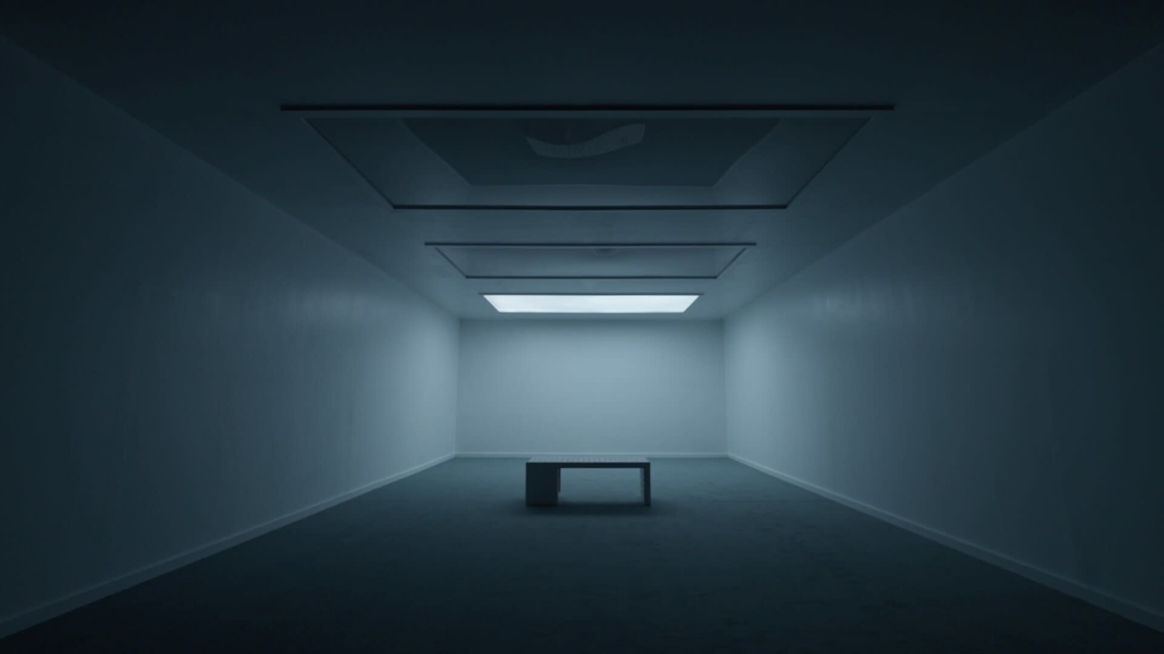 A sparsely furnished room is illuminated by a skylight, featuring a single table in the center and plain walls.