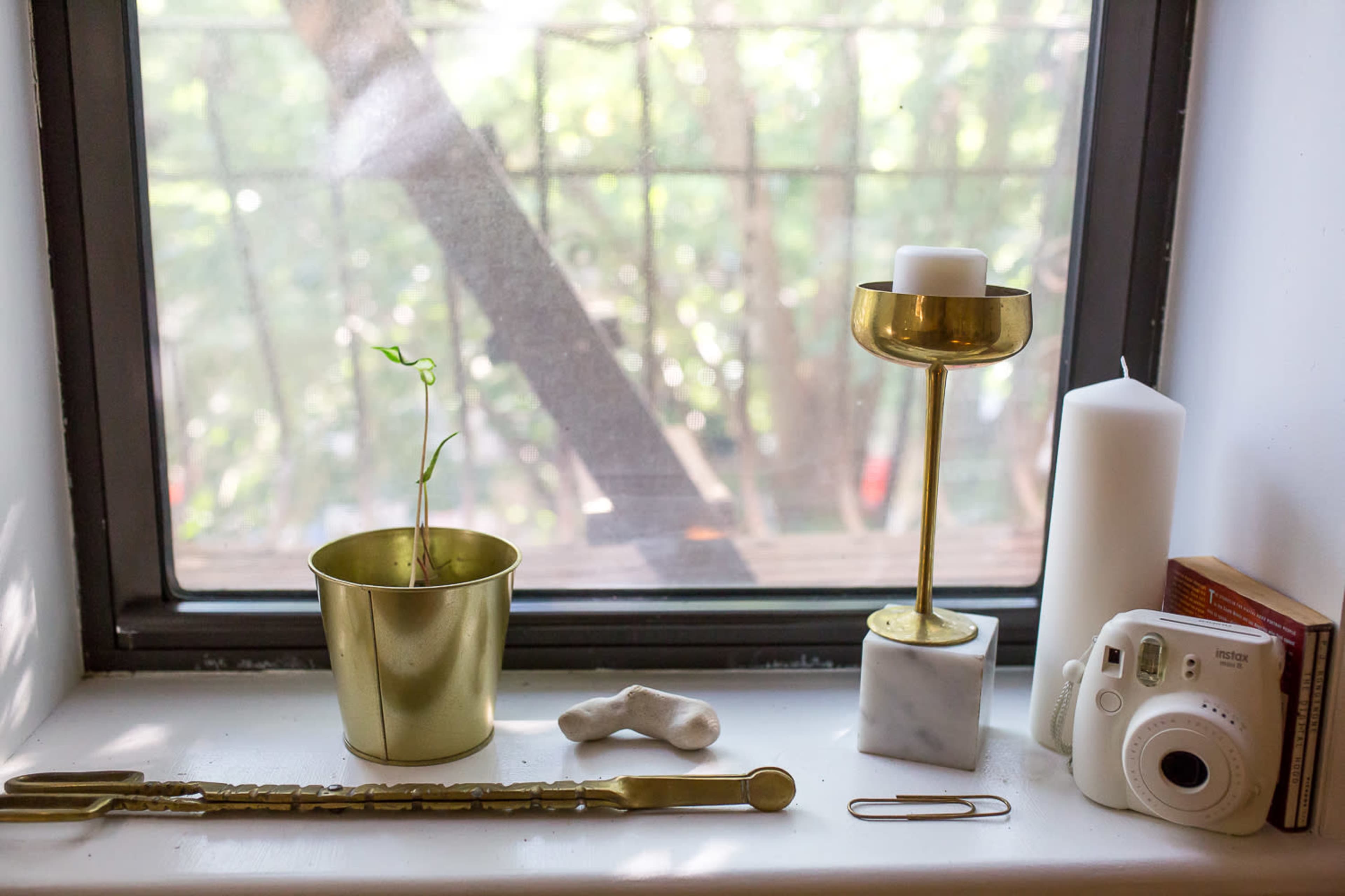 A small brass pot with a green plant, a gold candle holder with a white candle, a white candle, a marble trinket, a clay object, a paperclip, and a camera are arranged on a windowsill with a view of trees outside.