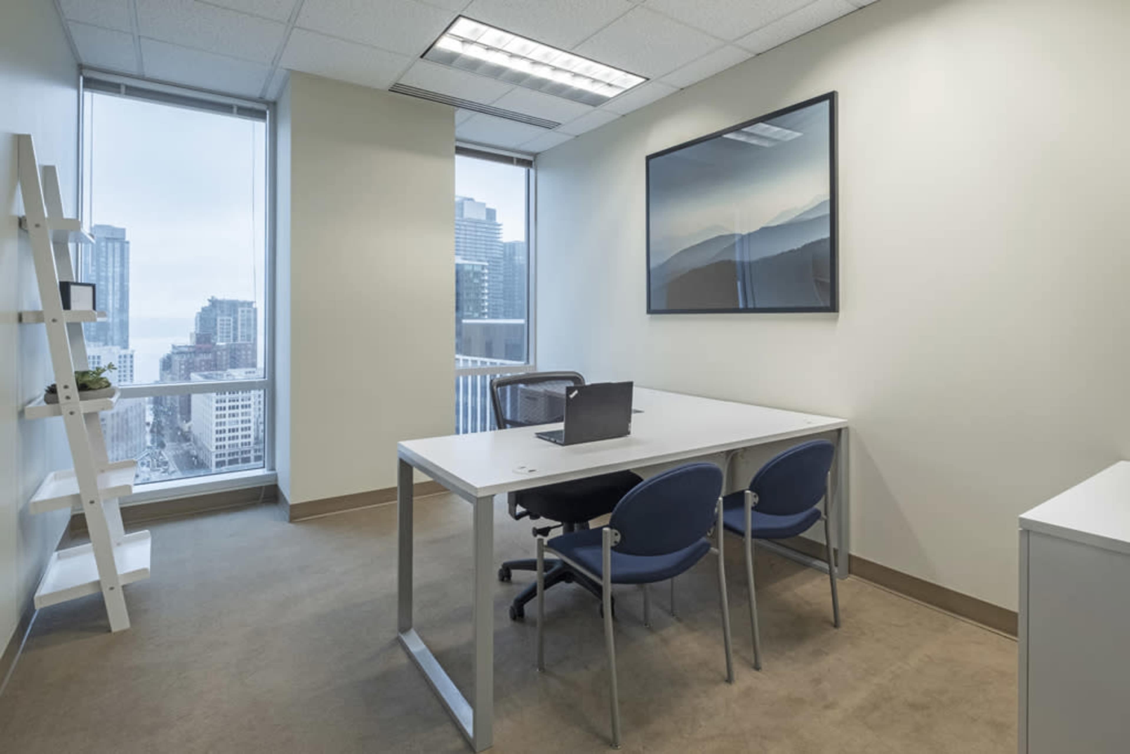 The image shows a modern office space featuring a white desk with a laptop, two blue chairs, and large windows overlooking a cityscape.