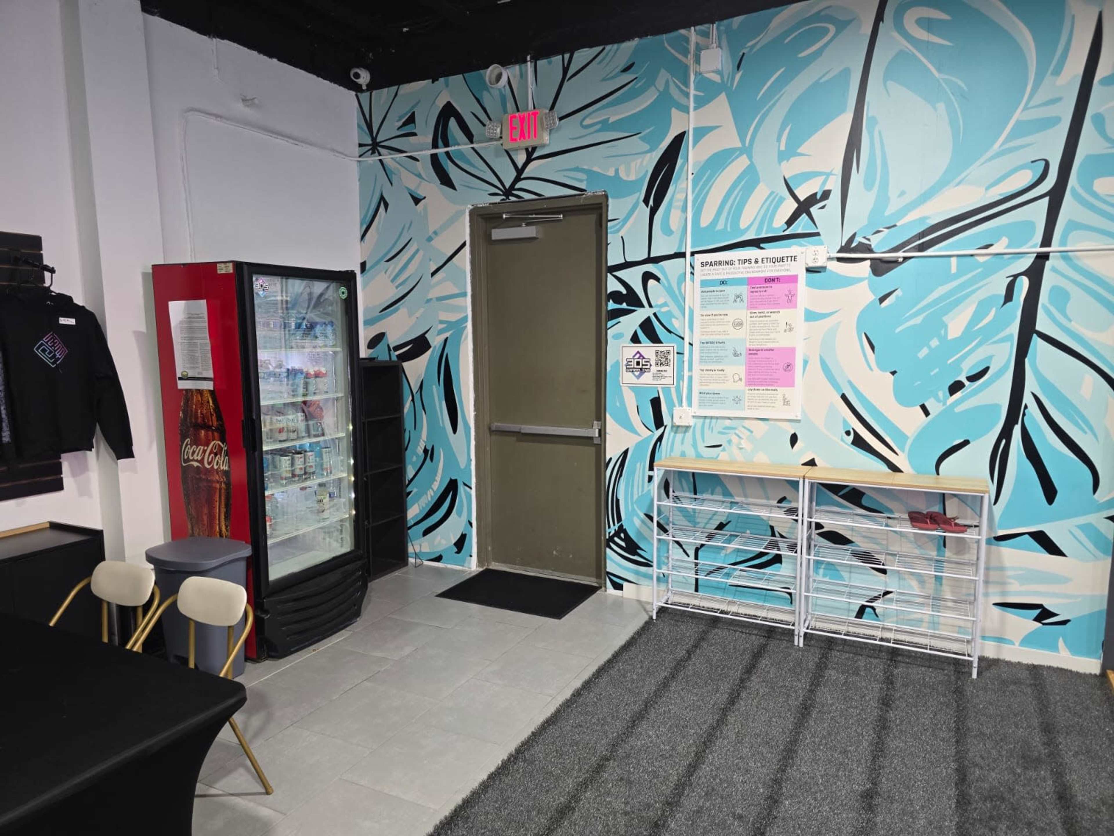 The image shows a small interior space featuring a blue mural on the wall, a door, a Coca-Cola vending machine, and a notice board with informational posters.