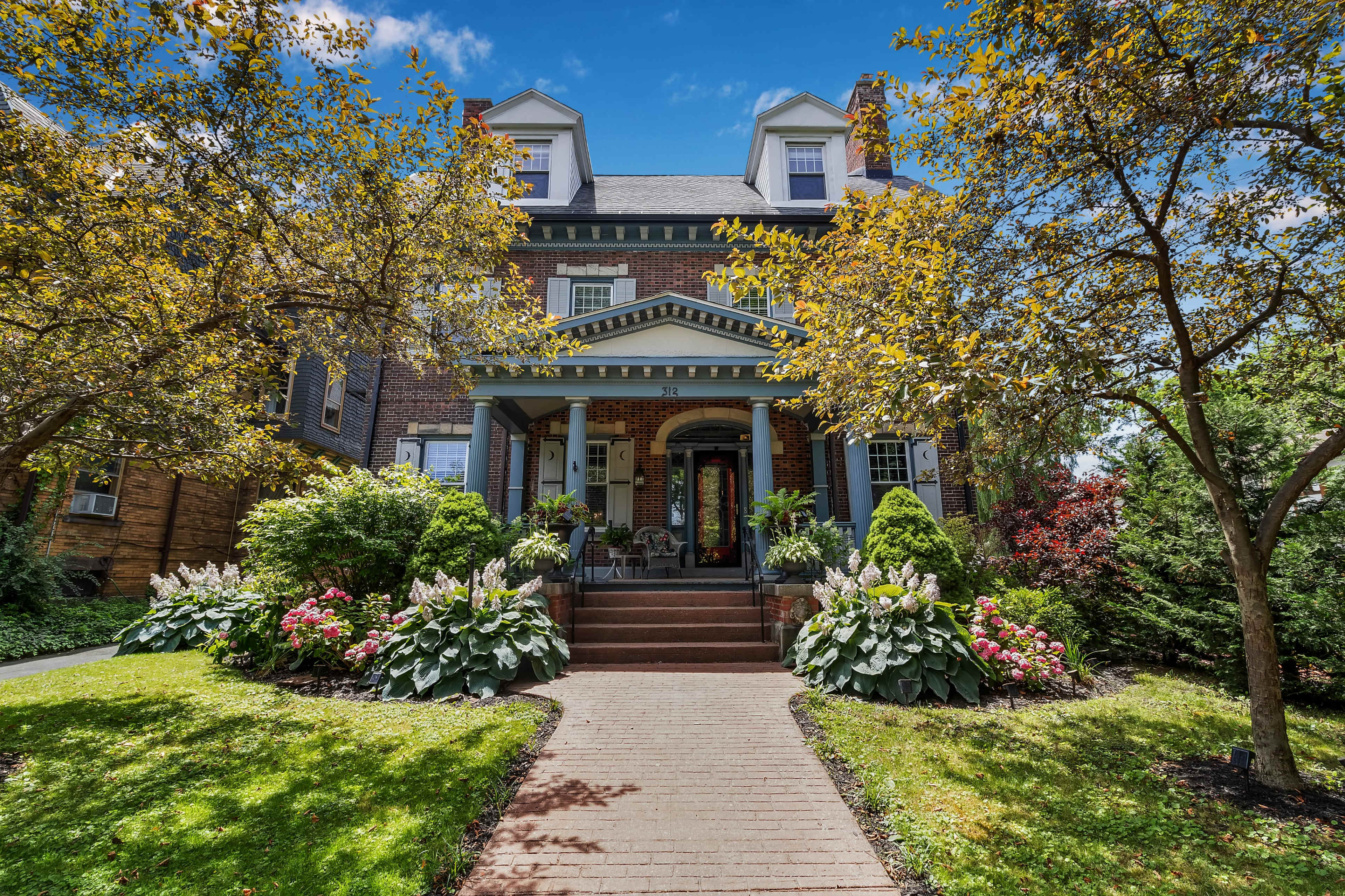 A large brick house with a central entrance and well-maintained gardens featuring colorful flowering plants and trees.