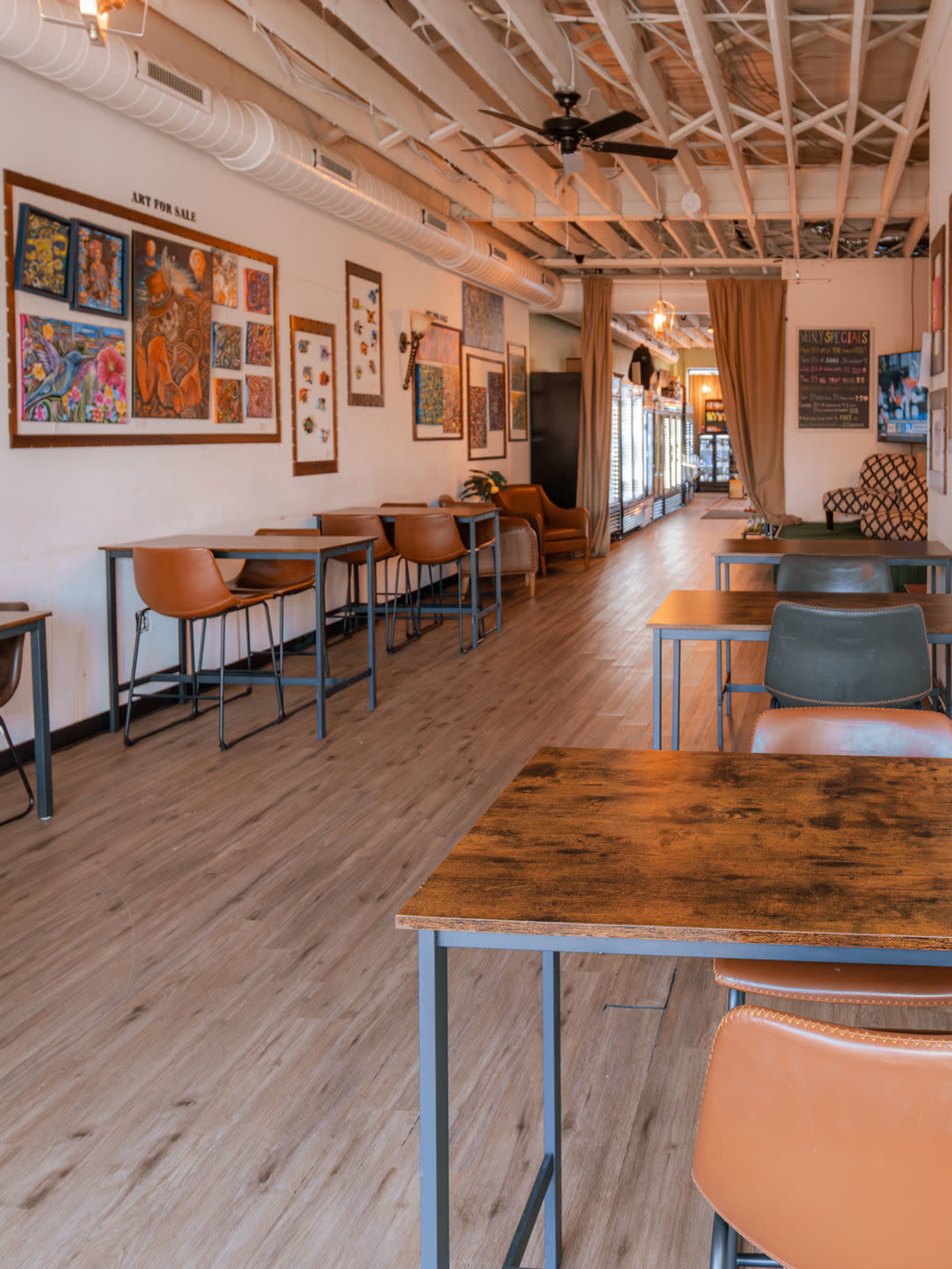 The image shows a spacious café with wooden flooring, rows of tables and chairs, and walls decorated with colorful artwork.