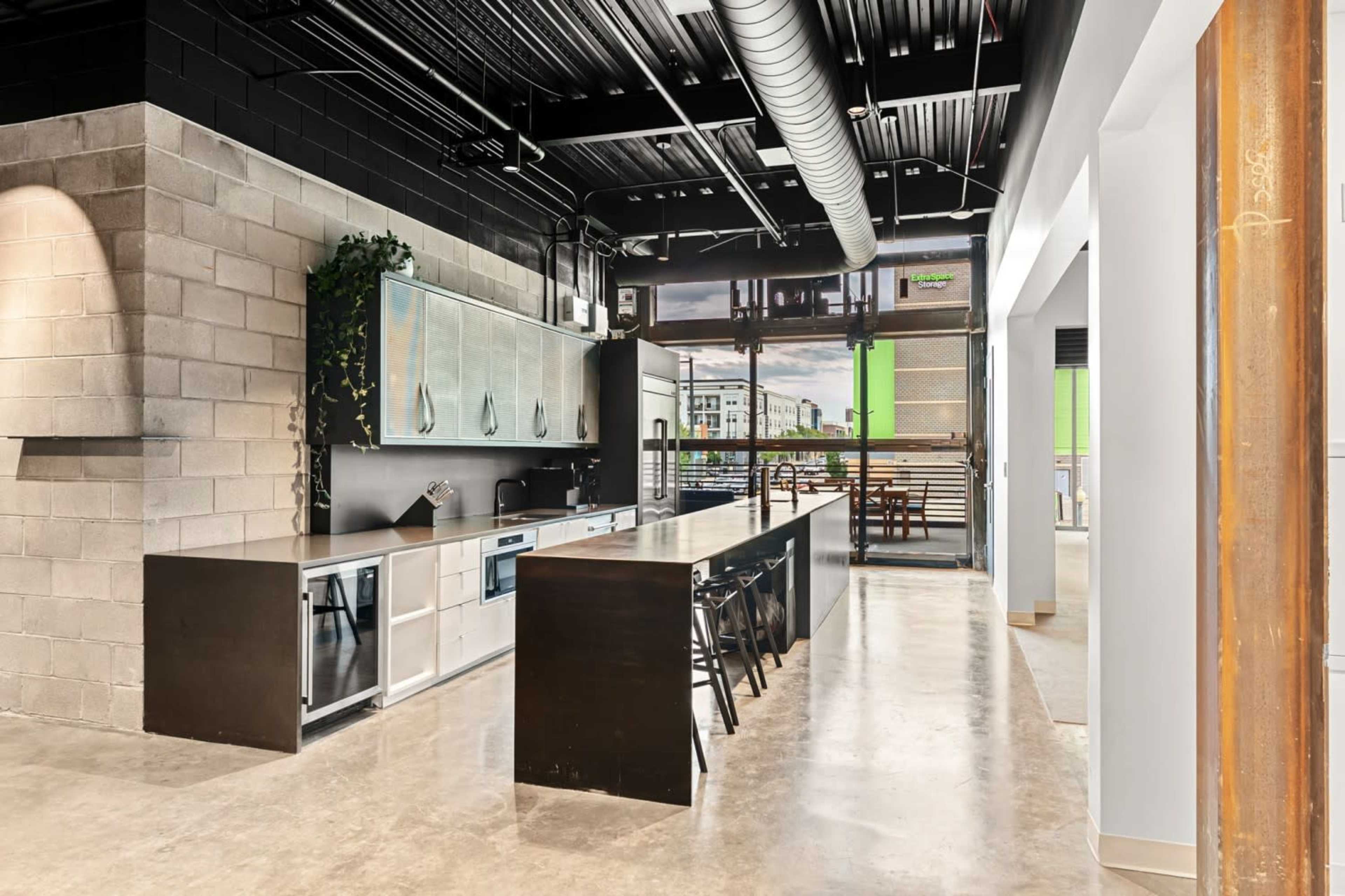 The image shows a modern kitchen with a black island, concrete flooring, and large windows that provide natural light.