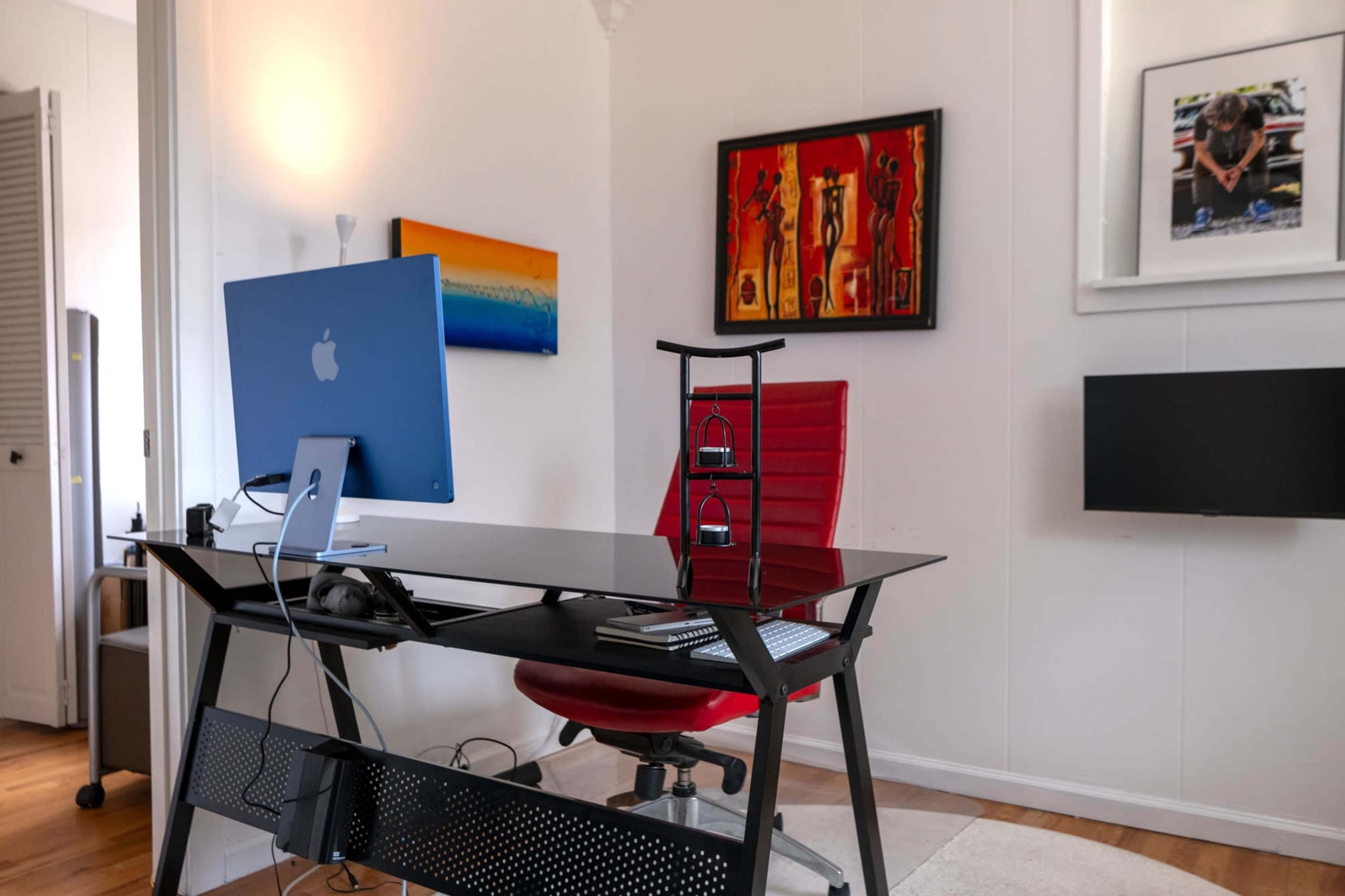 The image shows a contemporary home office featuring a black desk with an iMac, a red ergonomic chair, and colorful wall art.