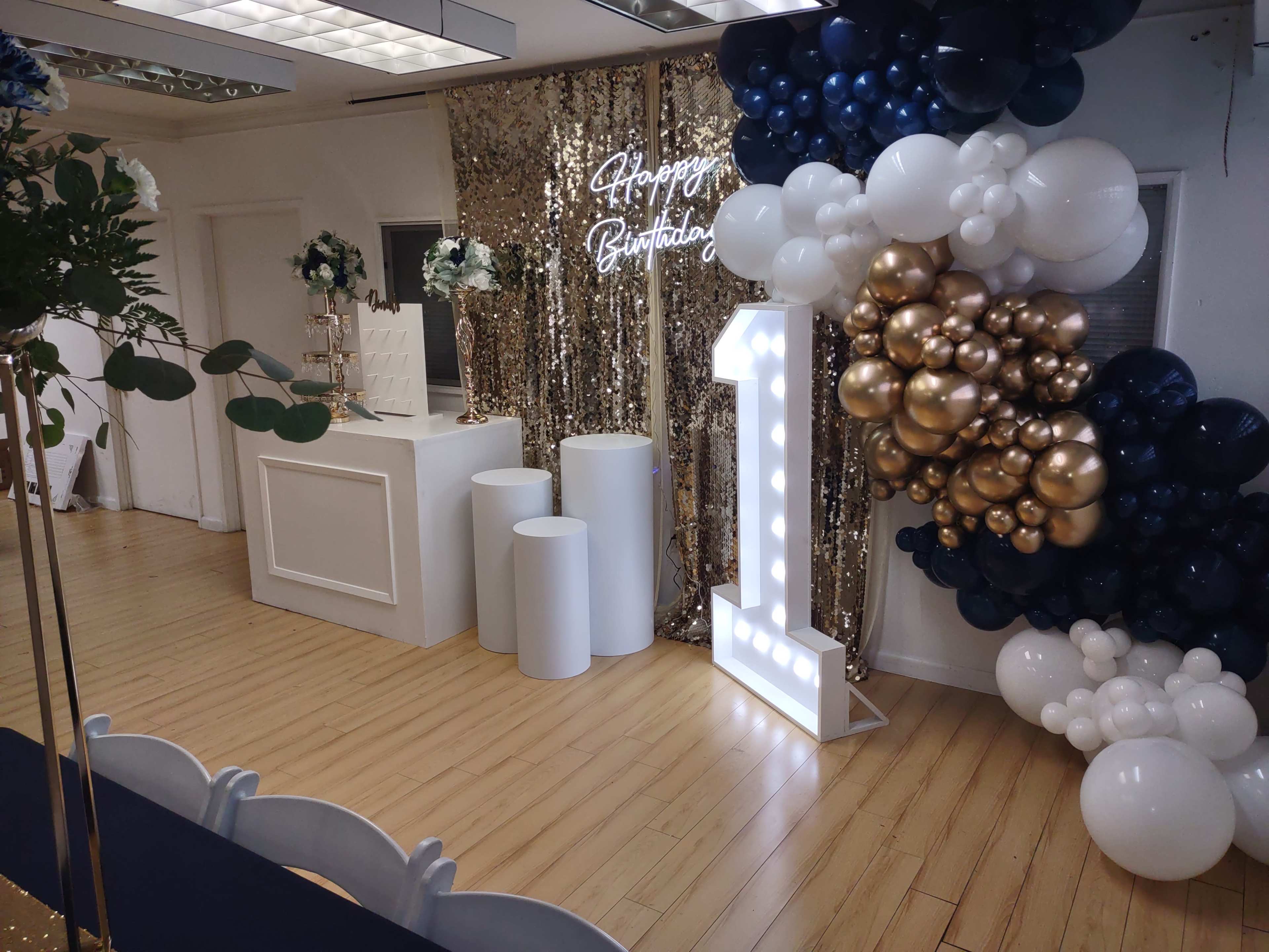 A decorated event space with a "Happy Birthday" sign, a large illuminated number one, and a backdrop of balloons and sequin fabric.