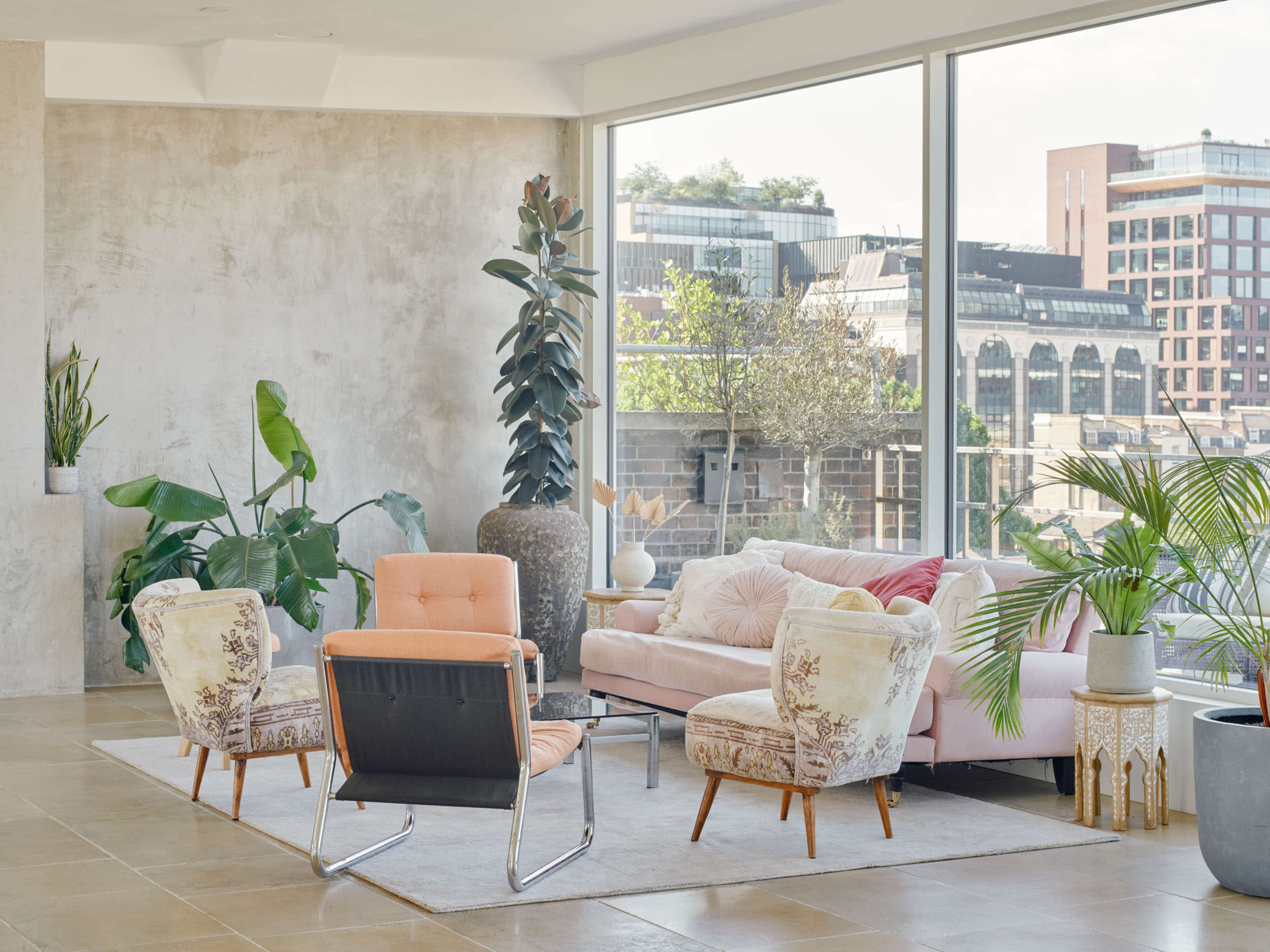 The image features a modern living room with a pink sofa, patterned chairs, and large windows showcasing city views, surrounded by indoor plants.