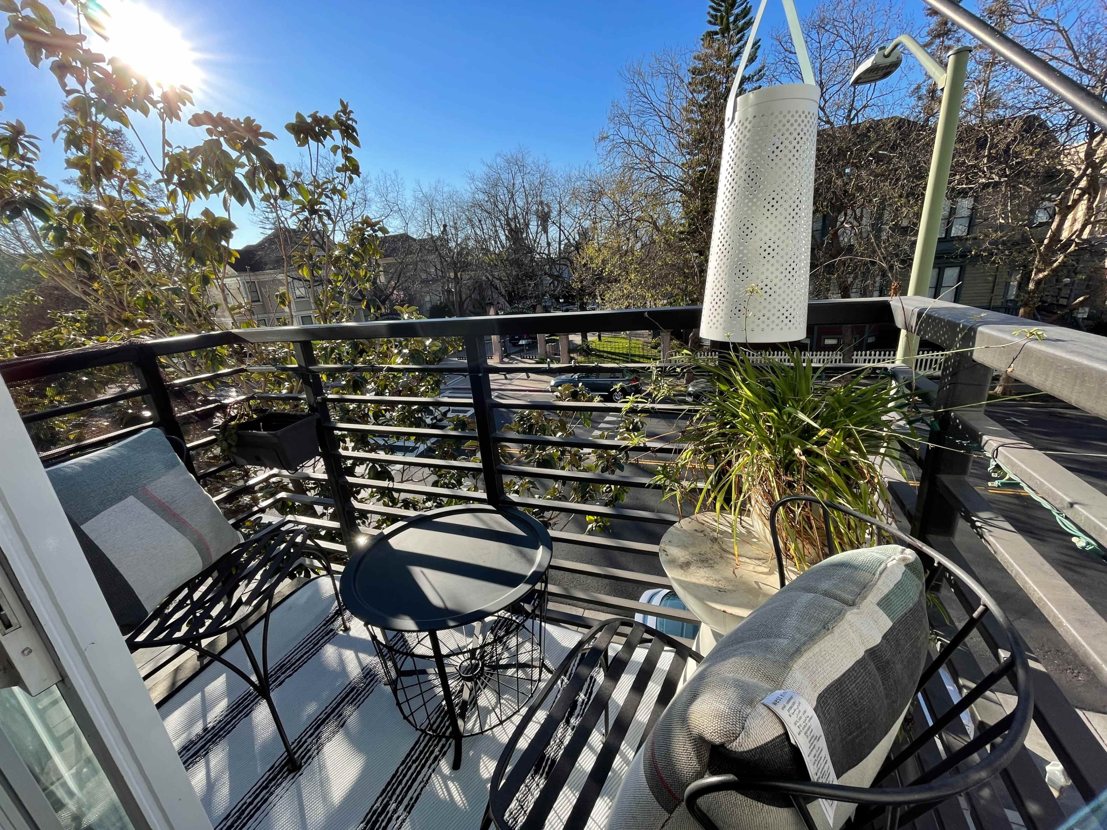 A balcony features two chairs and a small table, surrounded by greenery and overlooking a street.