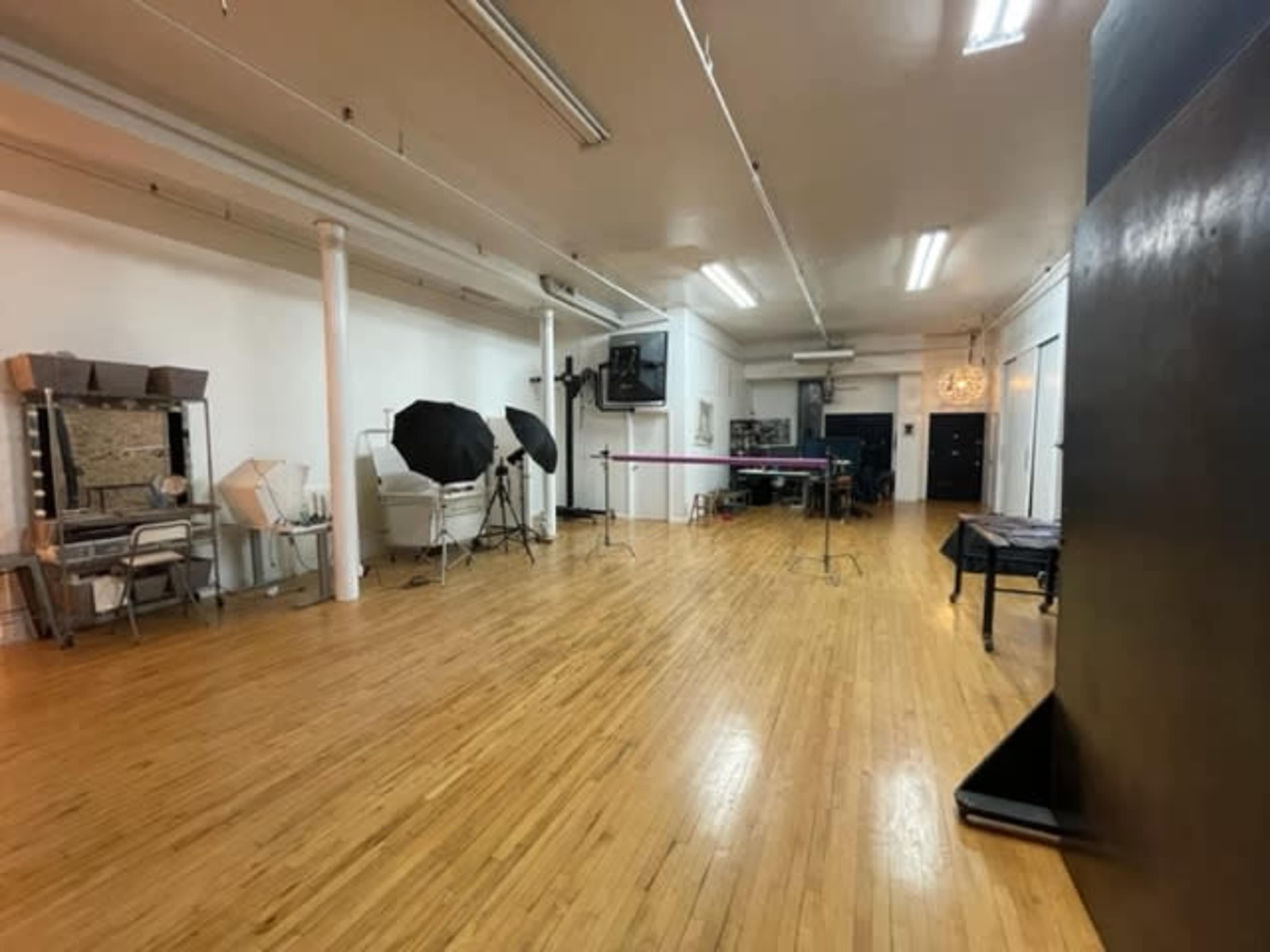 The image shows a spacious studio with wooden floors, featuring mirrors, photography equipment, and a large black backdrop.