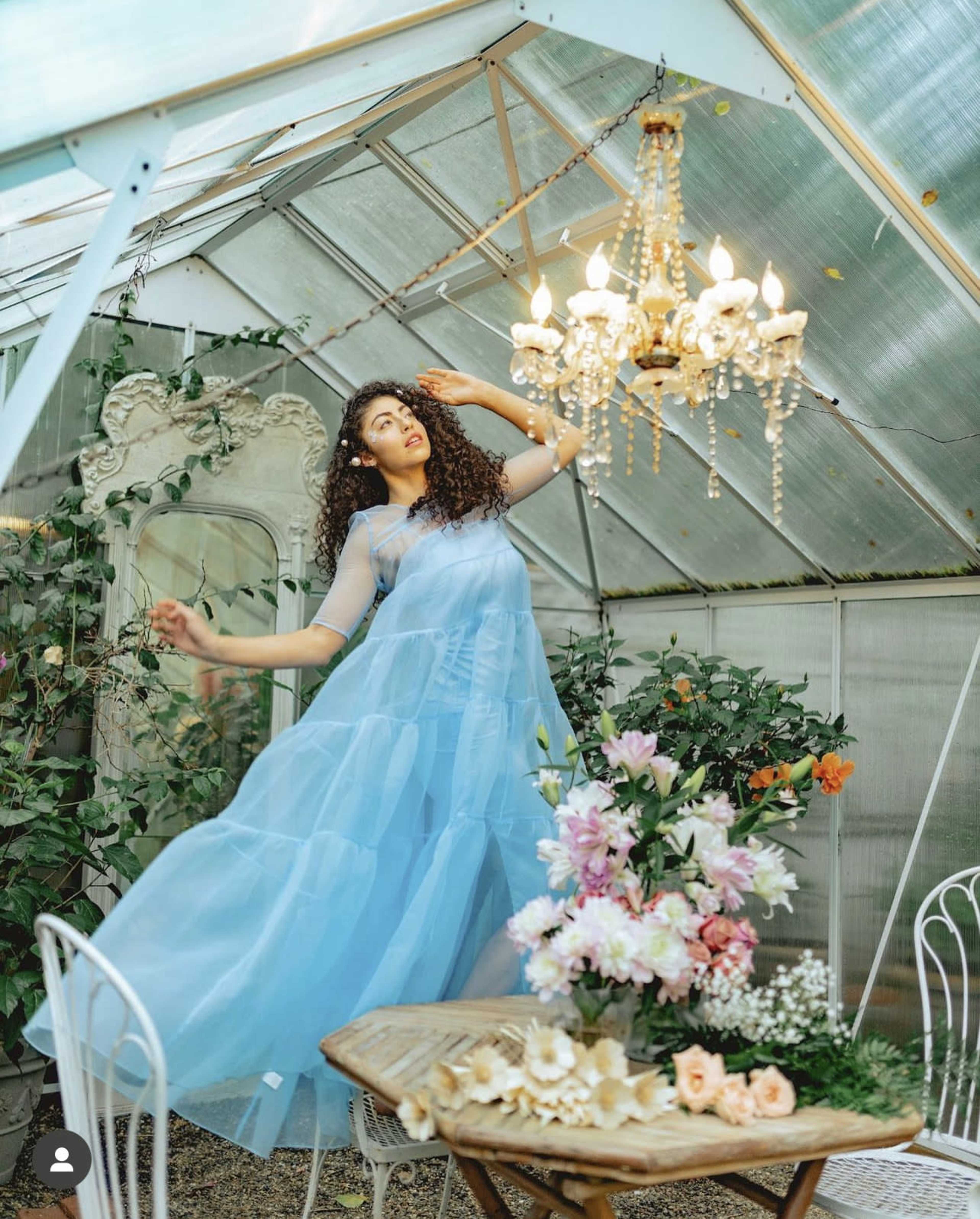 A woman in a flowing blue dress poses gracefully under a chandelier in a greenhouse filled with plants and a floral arrangement on a nearby table.
