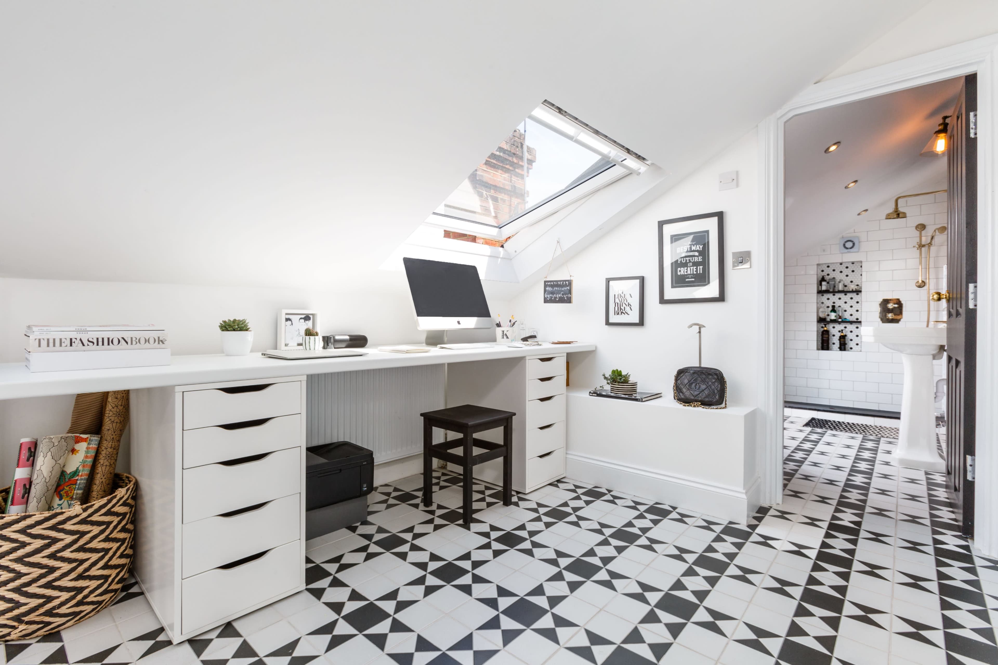 A modern attic office space with a desk and computer under a slanted ceiling, featuring patterned tile flooring and a small bathroom area visible through an open door.