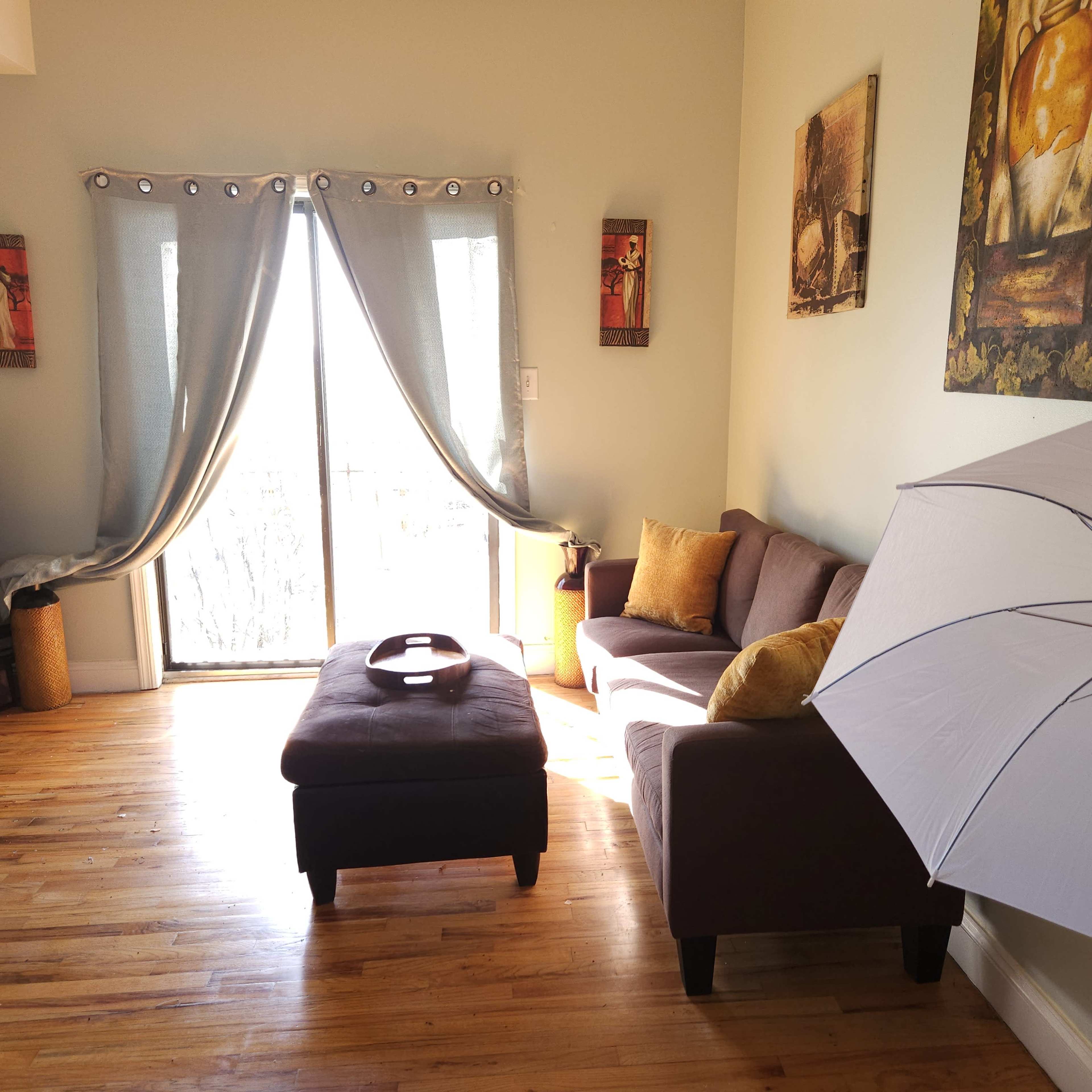 A living room features a brown couch, a dark ottoman, and large windows with sheer curtains letting in natural light.