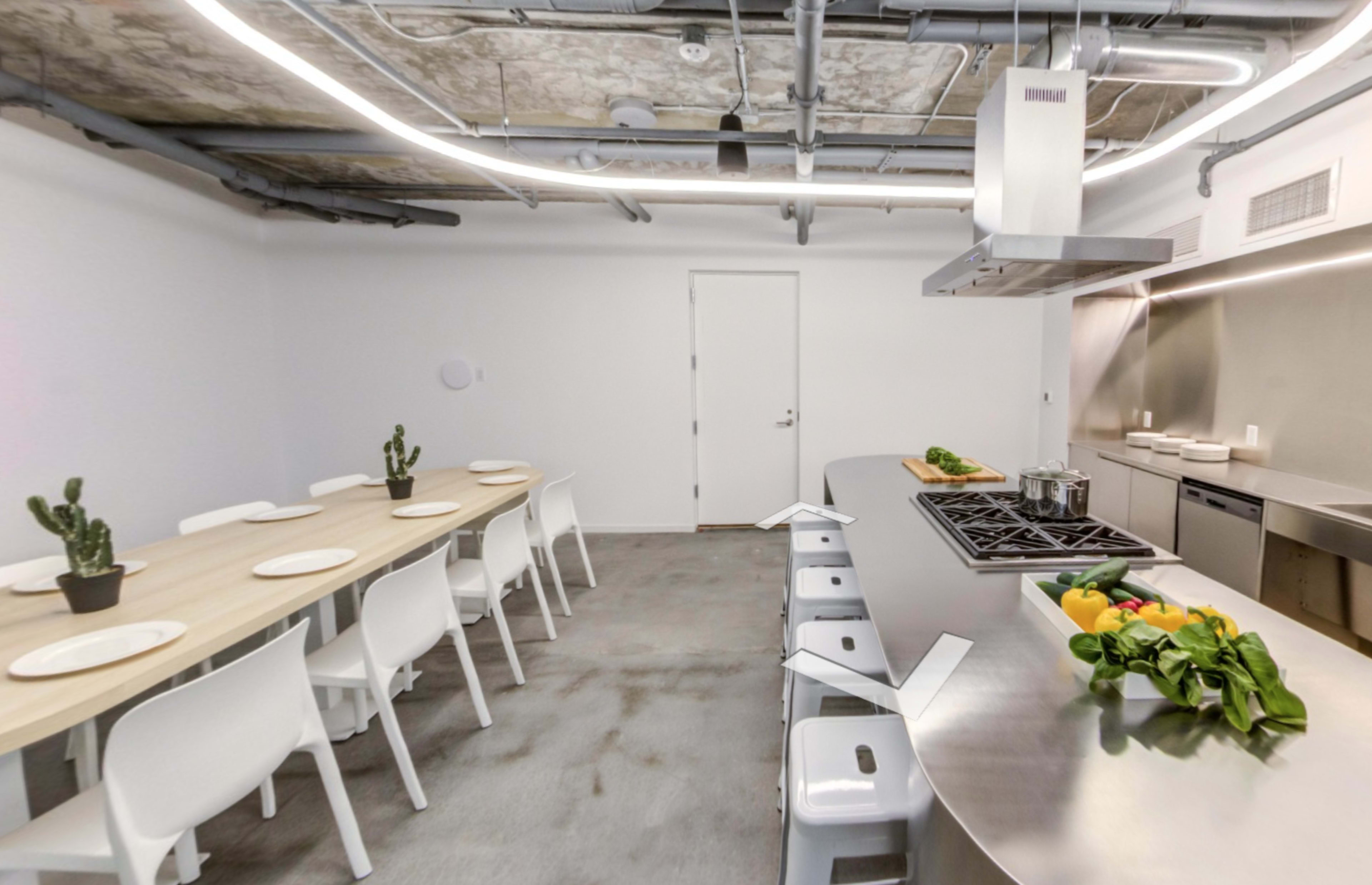 A modern kitchen features a long wooden table set for a meal, metal stools, and a stainless steel cooking area with a hood and stovetop.