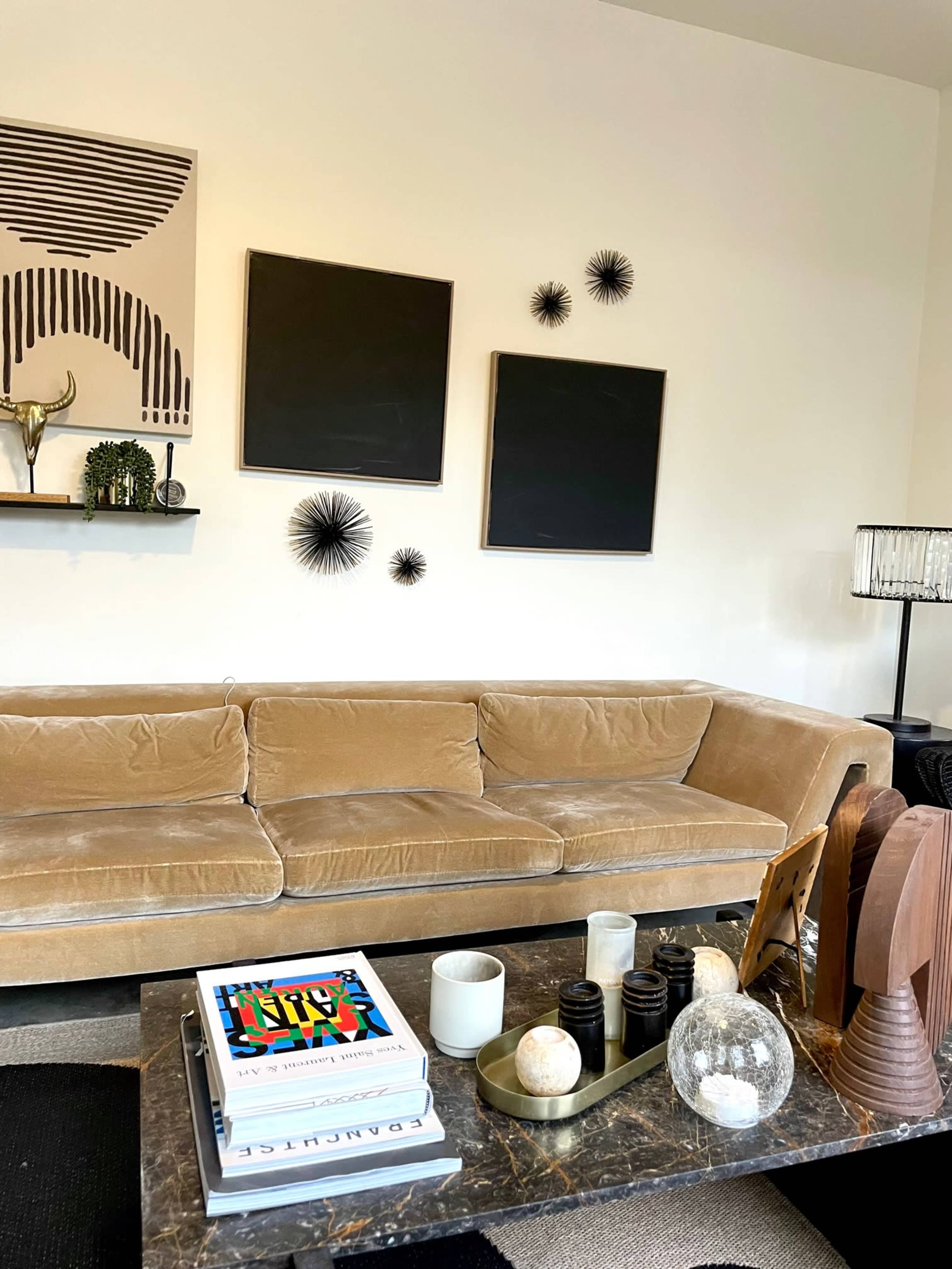 The image features a modern living room with a beige sofa, coffee table displaying various decorative items, and two black framed artworks on the wall.
