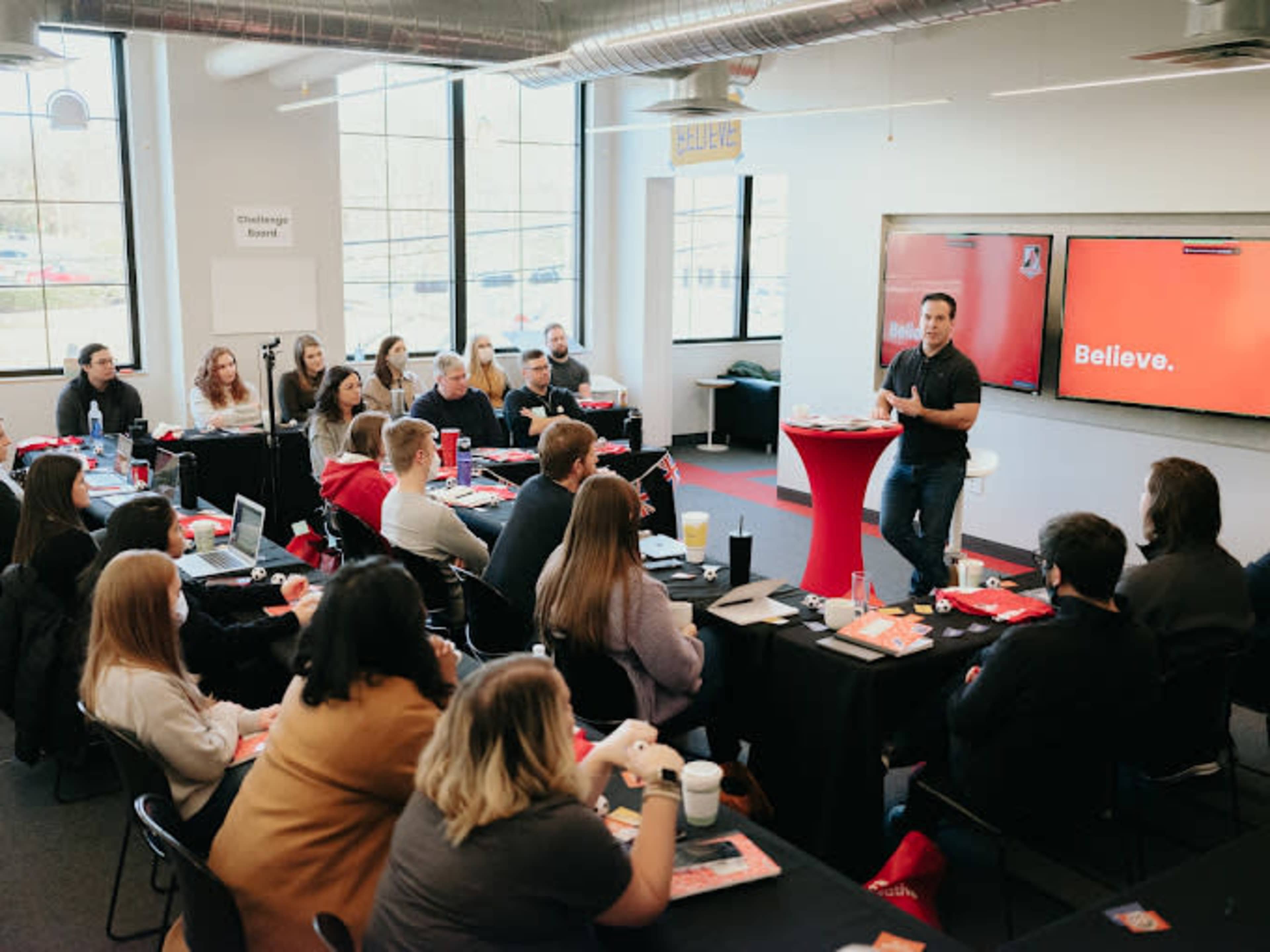 A speaker stands in front of an audience seated at tables in a conference room, presenting on a screen that displays the word "Believe."