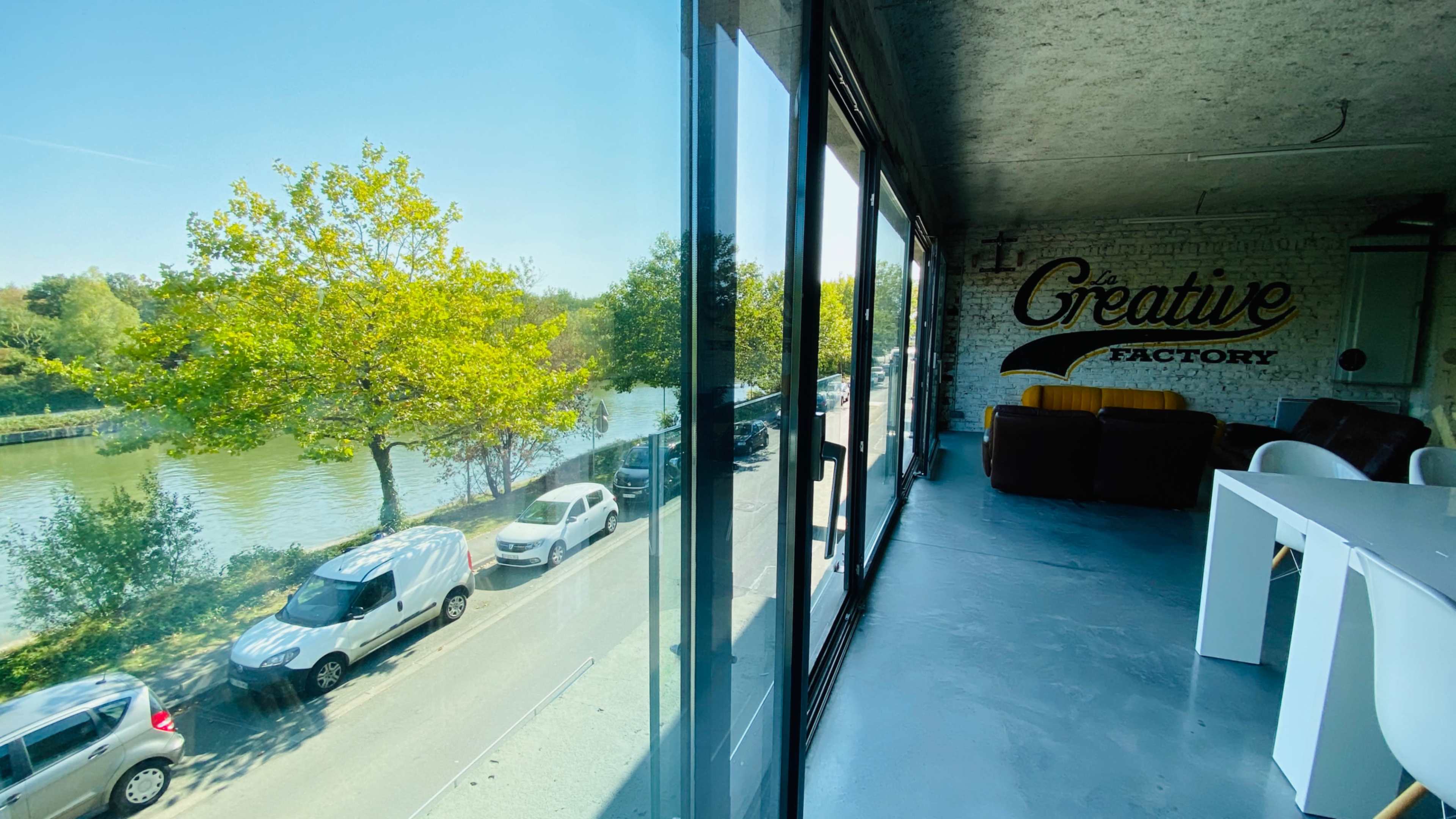 A modern workspace features large windows overlooking a tree-lined river and parked cars on a sunny day.