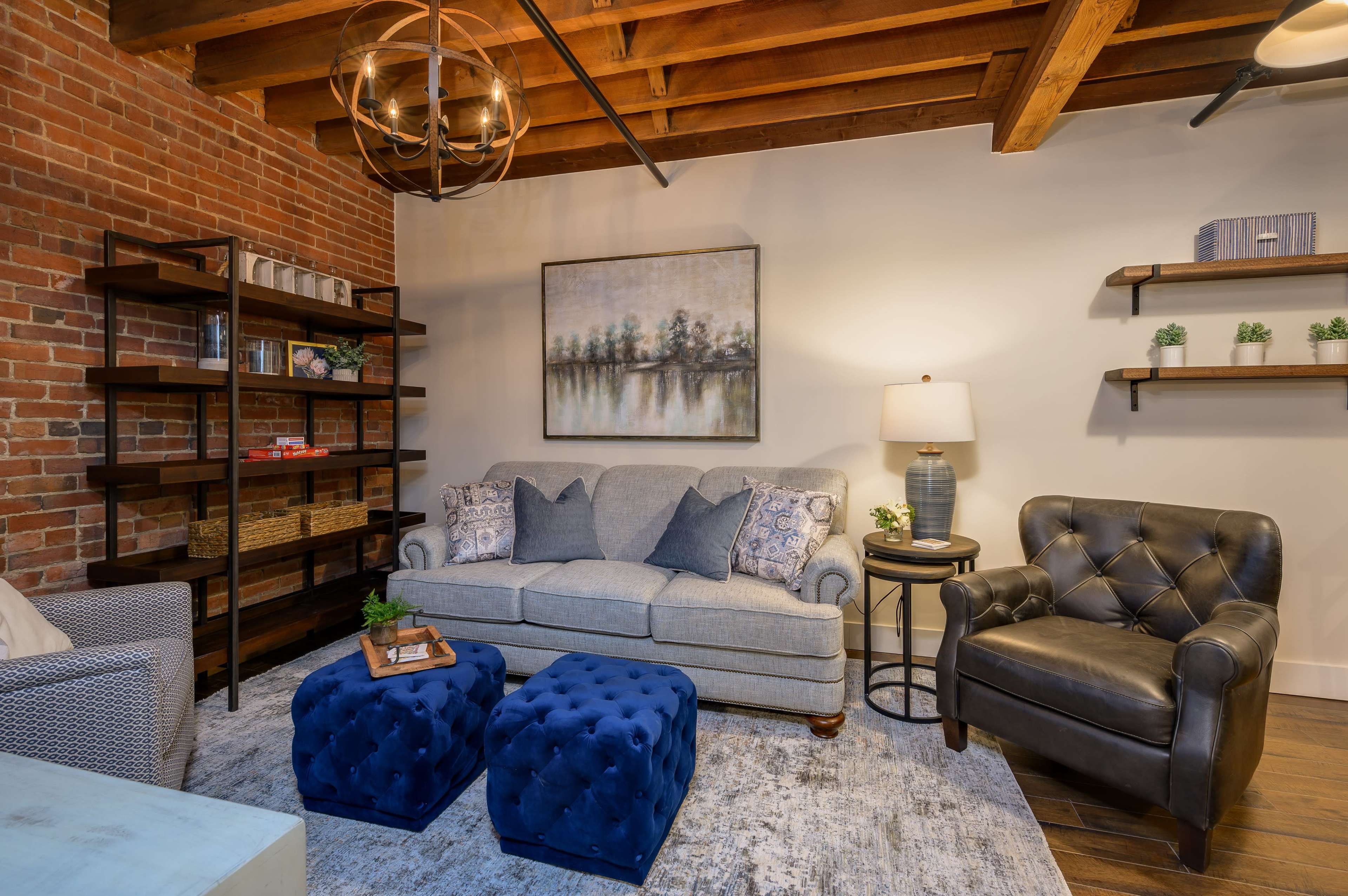 A cozy living room with a gray sofa, two blue tufted ottomans, a black leather armchair, and a brick wall adorned with shelves and artwork.