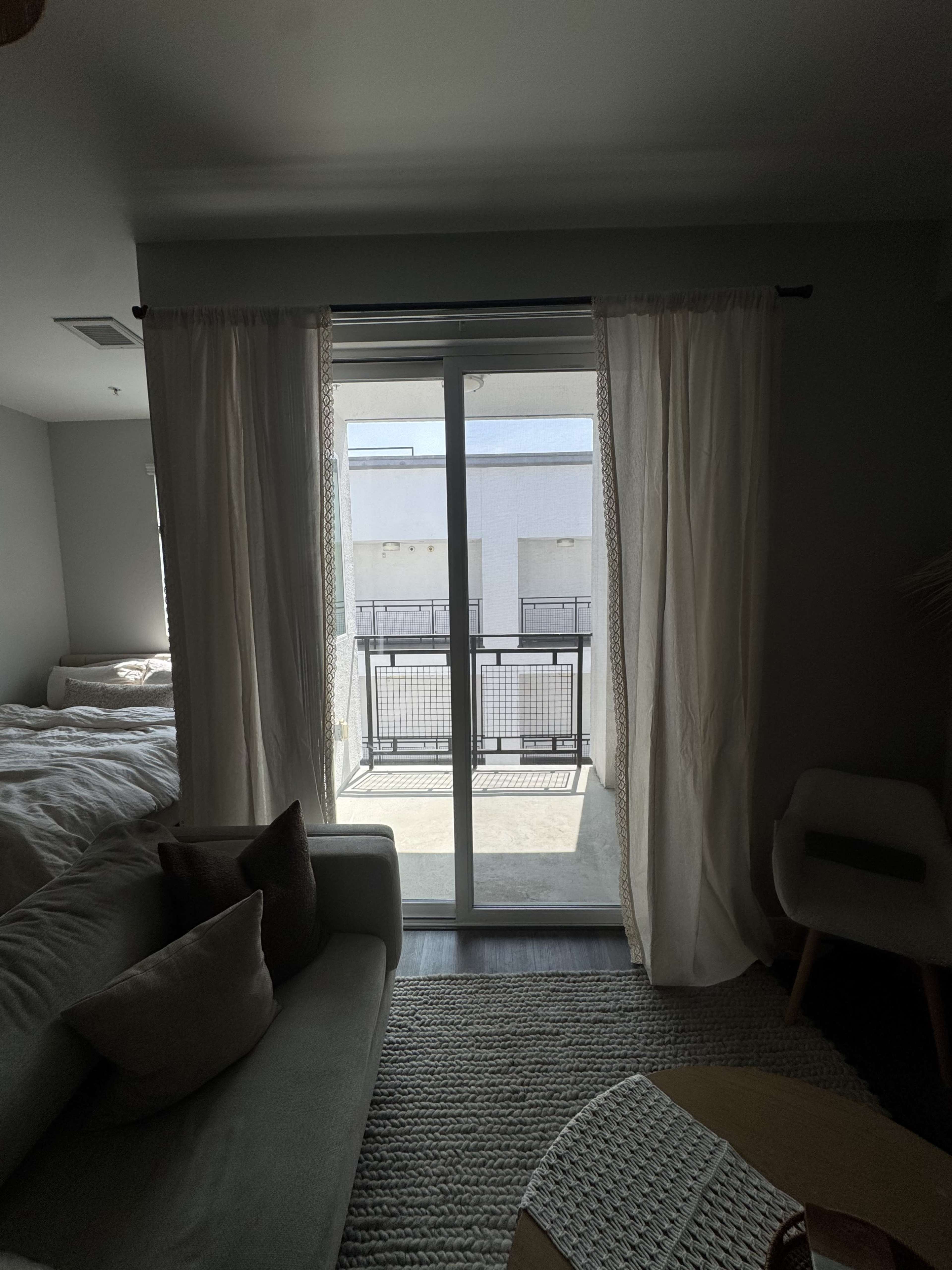 The image shows a room with a bed in the background, a couch with cushions in the foreground, and a glass door leading to a balcony.