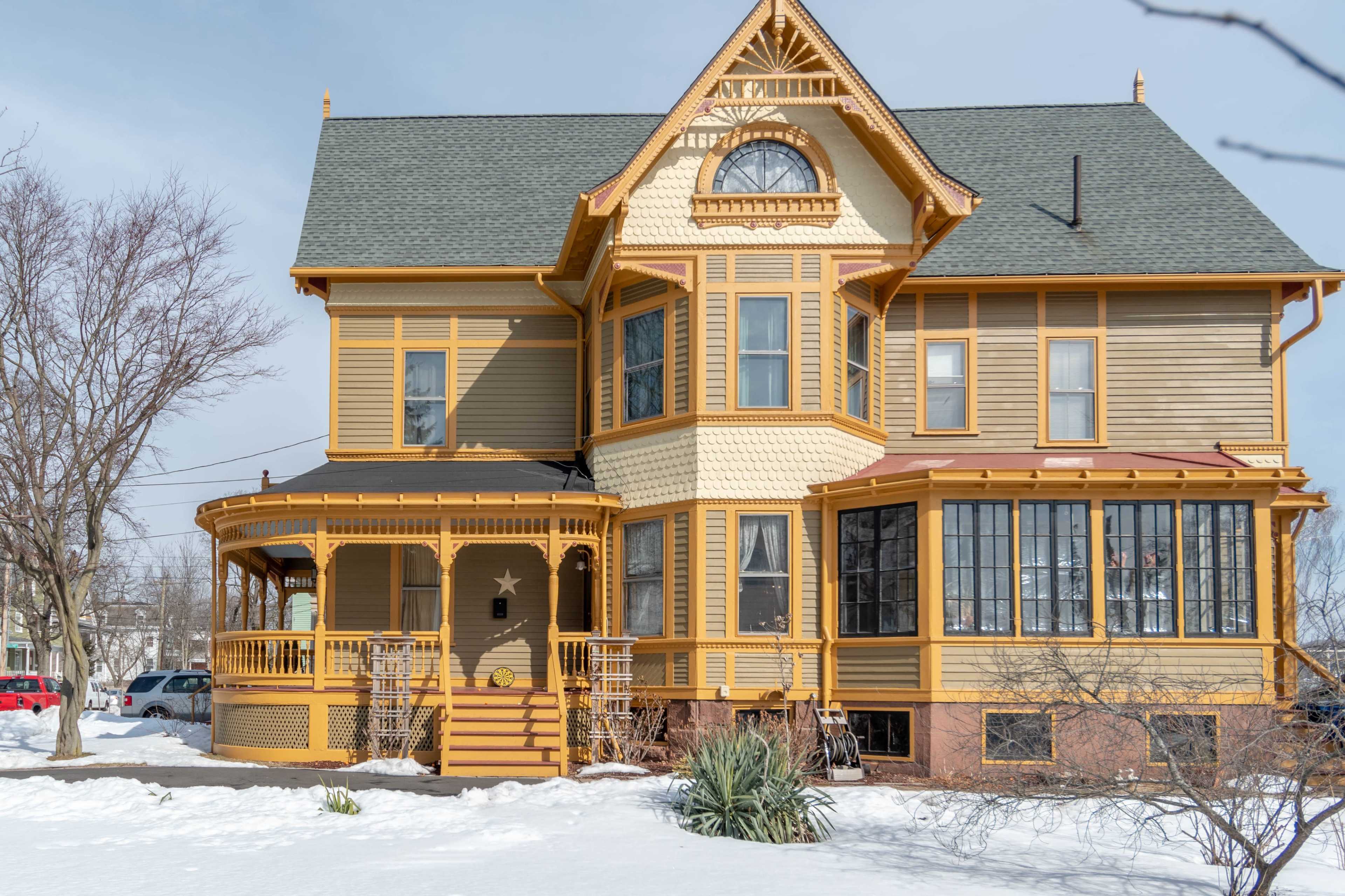 A large, two-story Victorian-style house with a wraparound porch and multiple windows is surrounded by snow.
