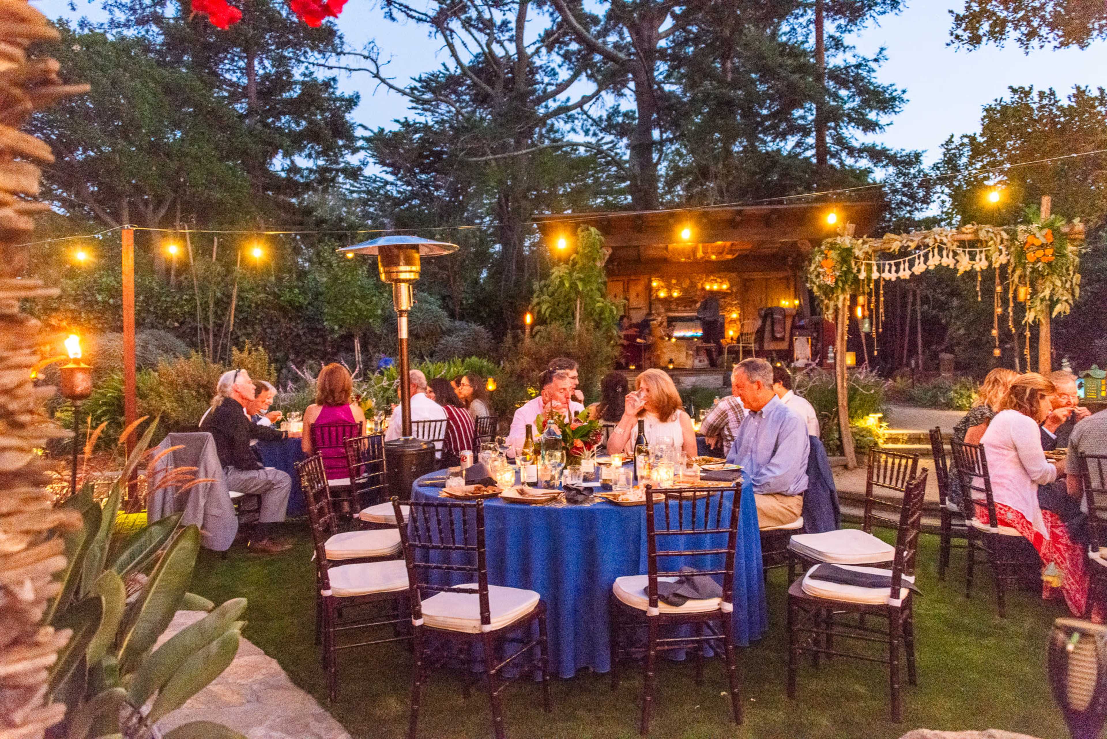 A garden dinner party is set up with tables and chairs, illuminated by string lights and surrounded by greenery.