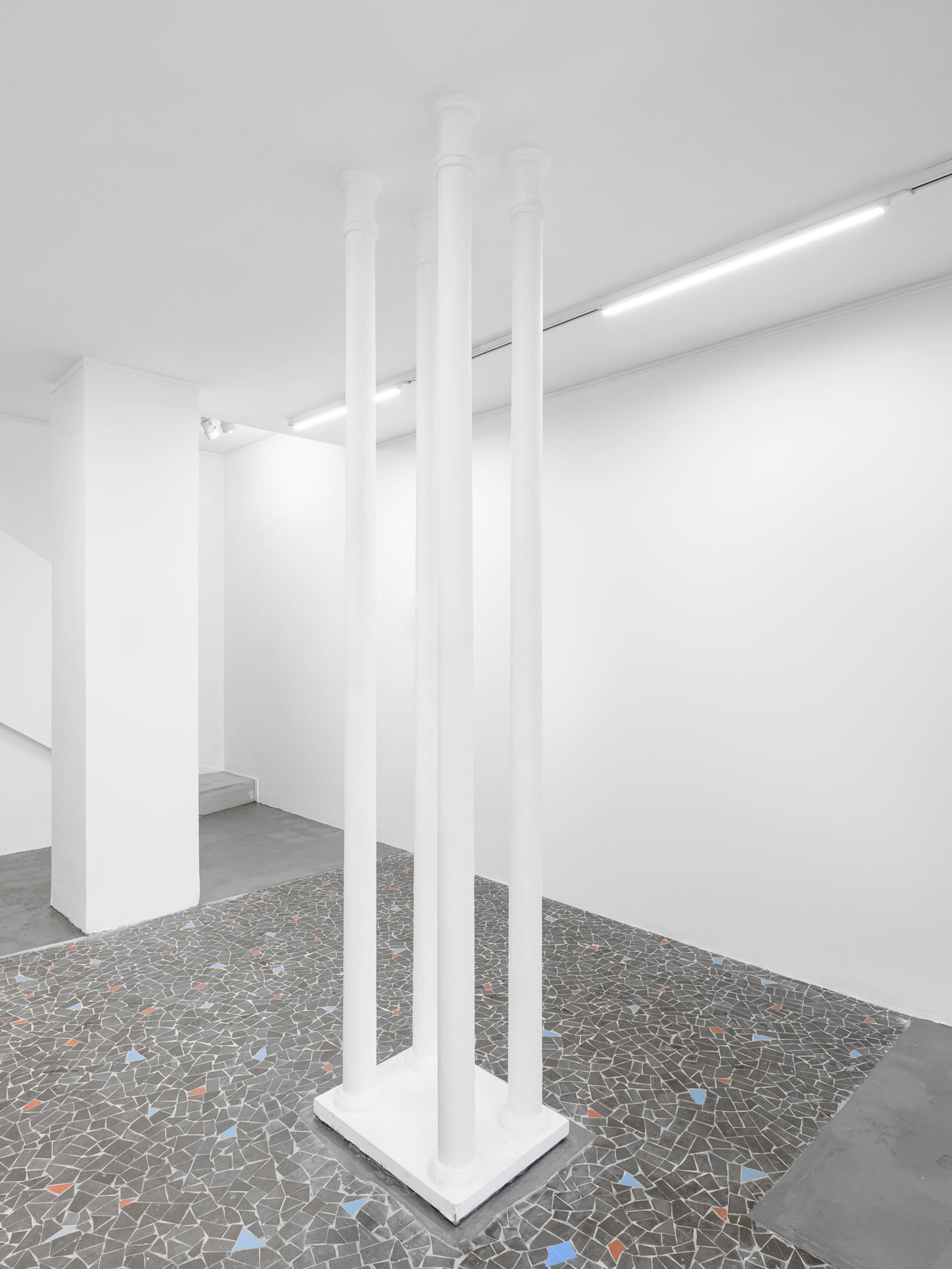 The image shows three white columns positioned on a tiled floor in a minimalist gallery space with white walls.