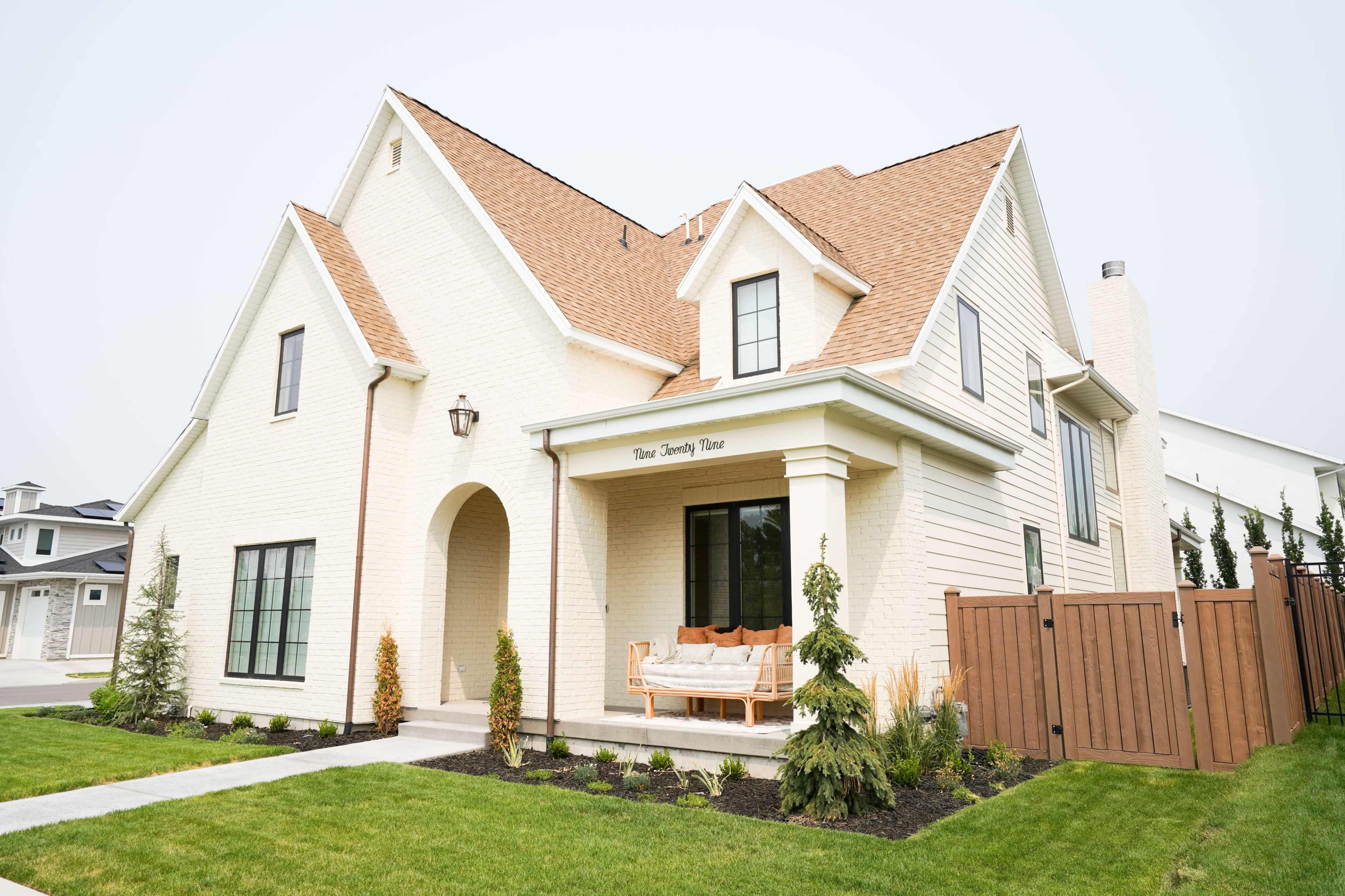 A two-story, light-colored house features a front porch with a bench, landscaped yard, and a wooden fence.
