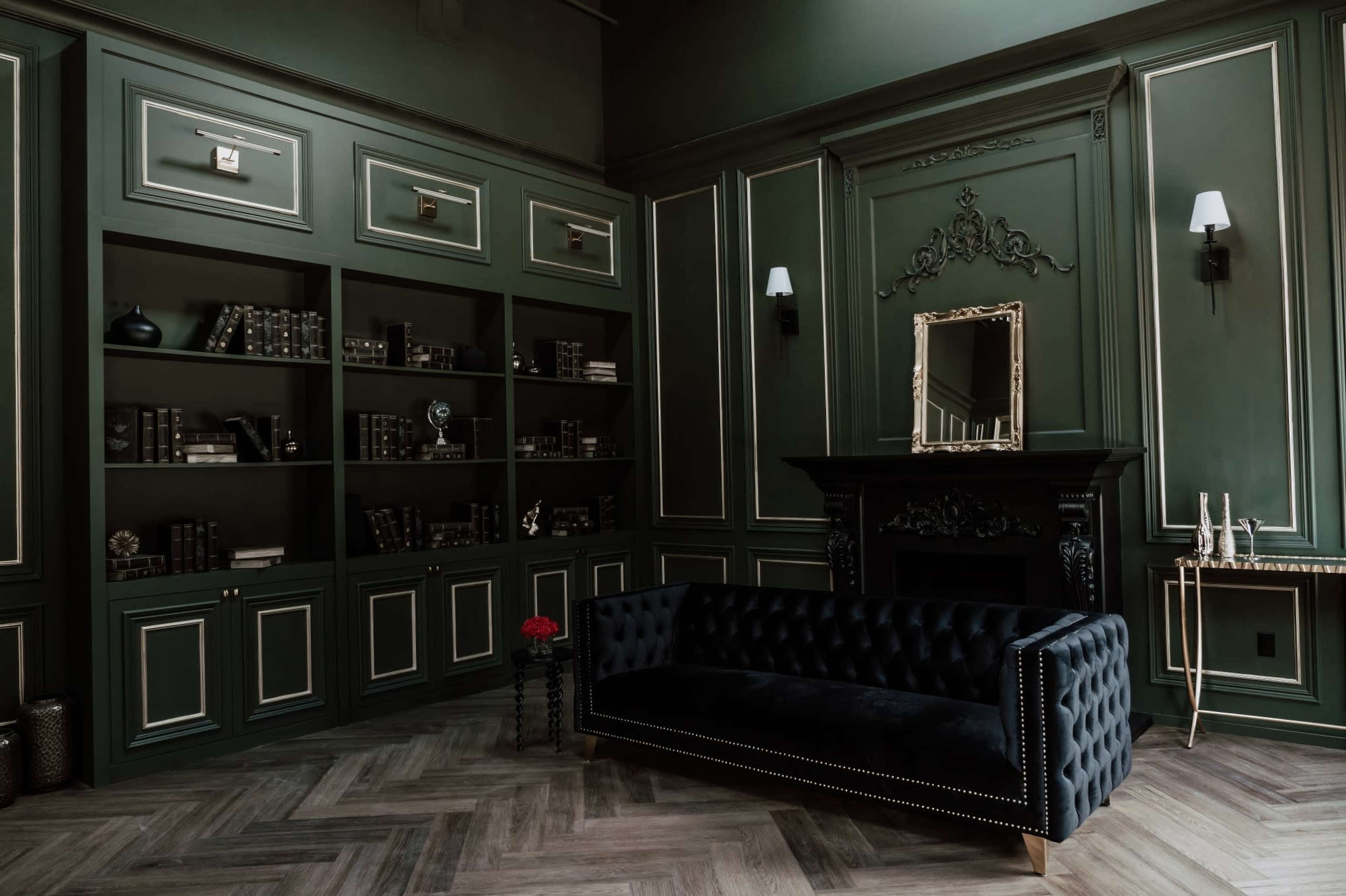 A room with dark green walls, a black tufted sofa, a fireplace, and a wall of bookshelves filled with books.