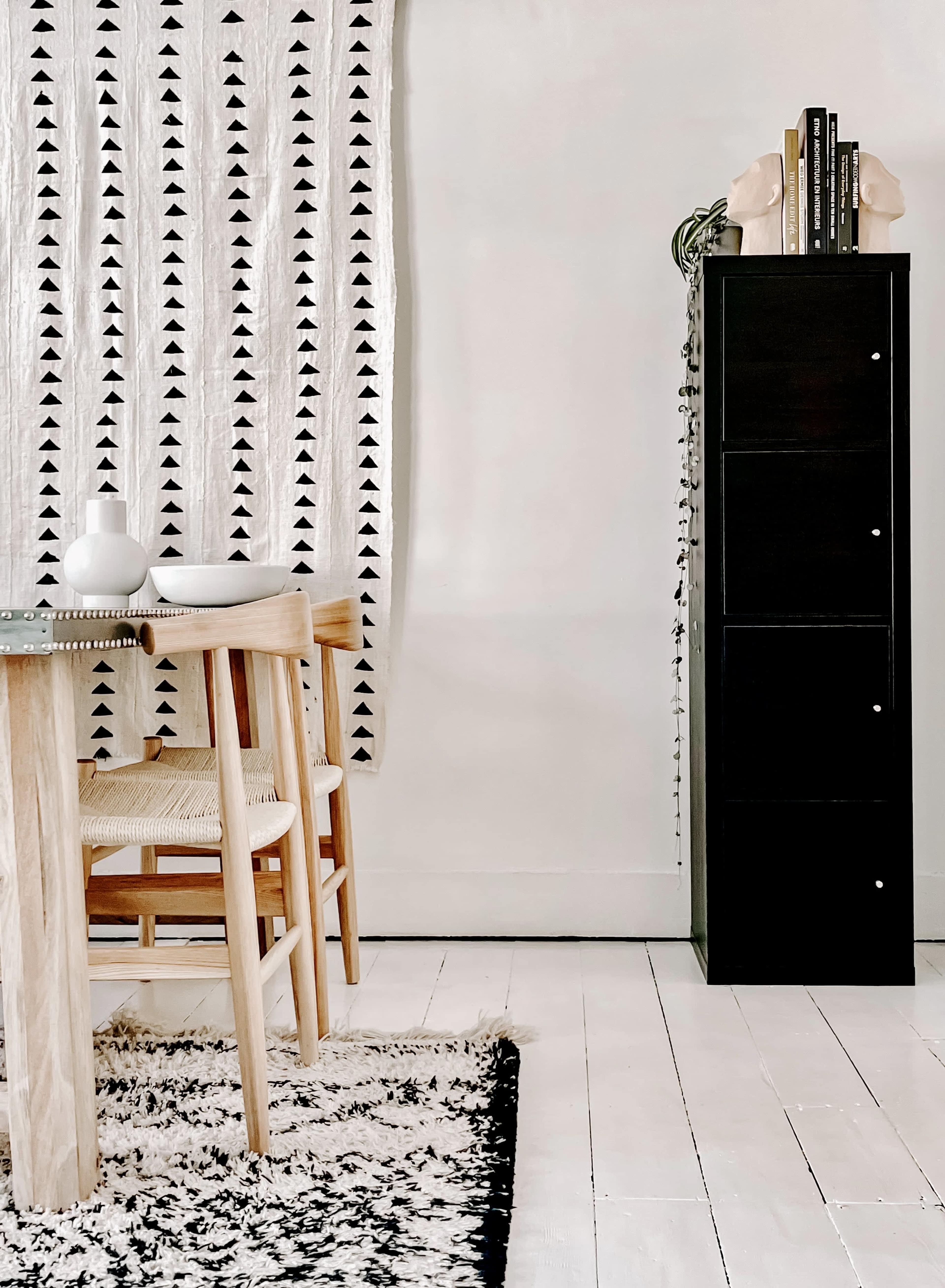 A minimalist interior featuring a wooden dining table with chairs, a patterned wall hanging, and a tall black storage cabinet against a light-colored wall.