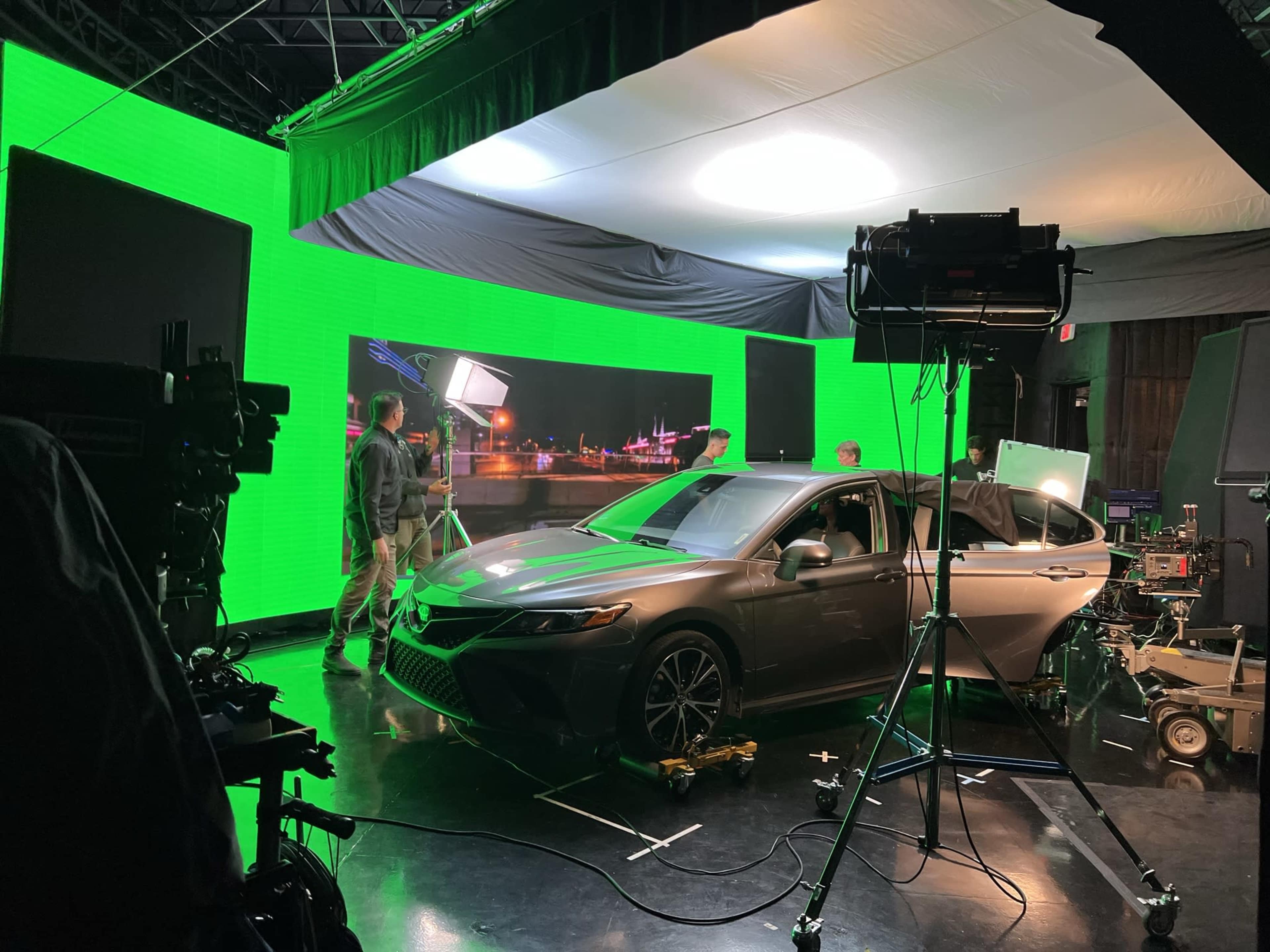 A film crew sets up a green screen studio with a car at the center, surrounded by lighting equipment and camera rigs.