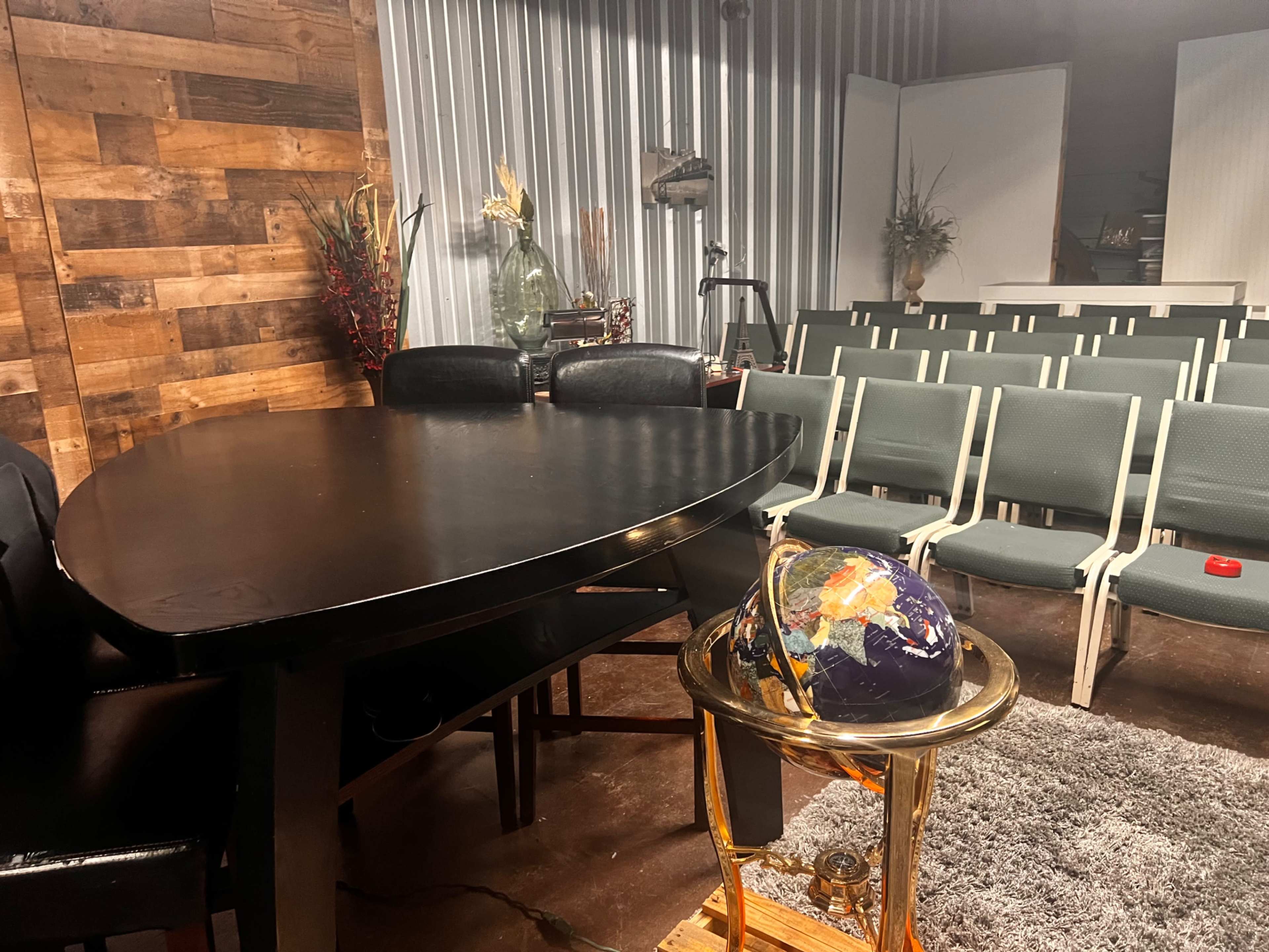 The scene shows a round black table and a globe on a gold stand, with a row of empty green chairs in a dimly lit room featuring wooden walls.