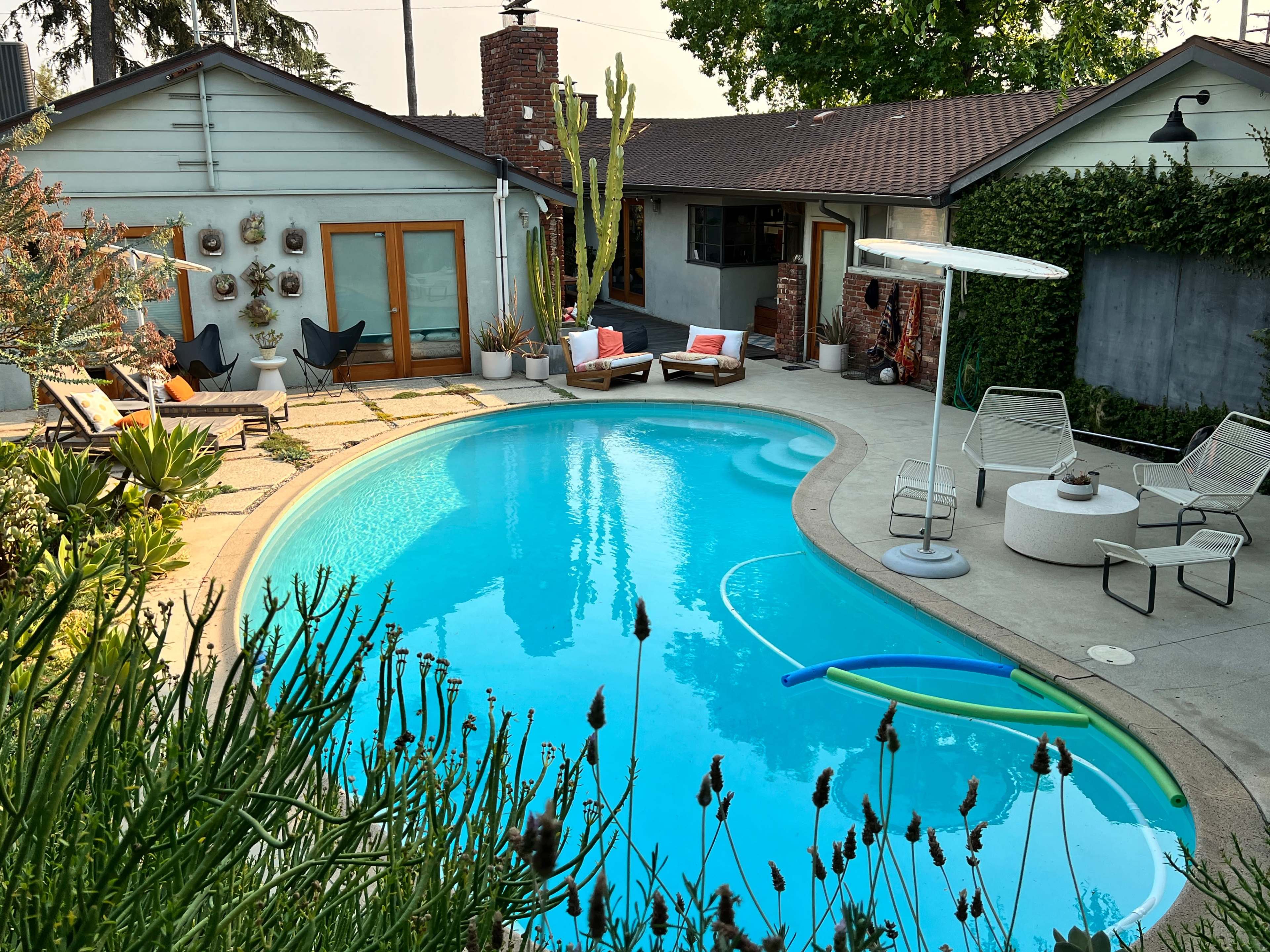 A backyard features a blue swimming pool surrounded by lounge chairs and patio furniture, with landscaping and a fence in the background.