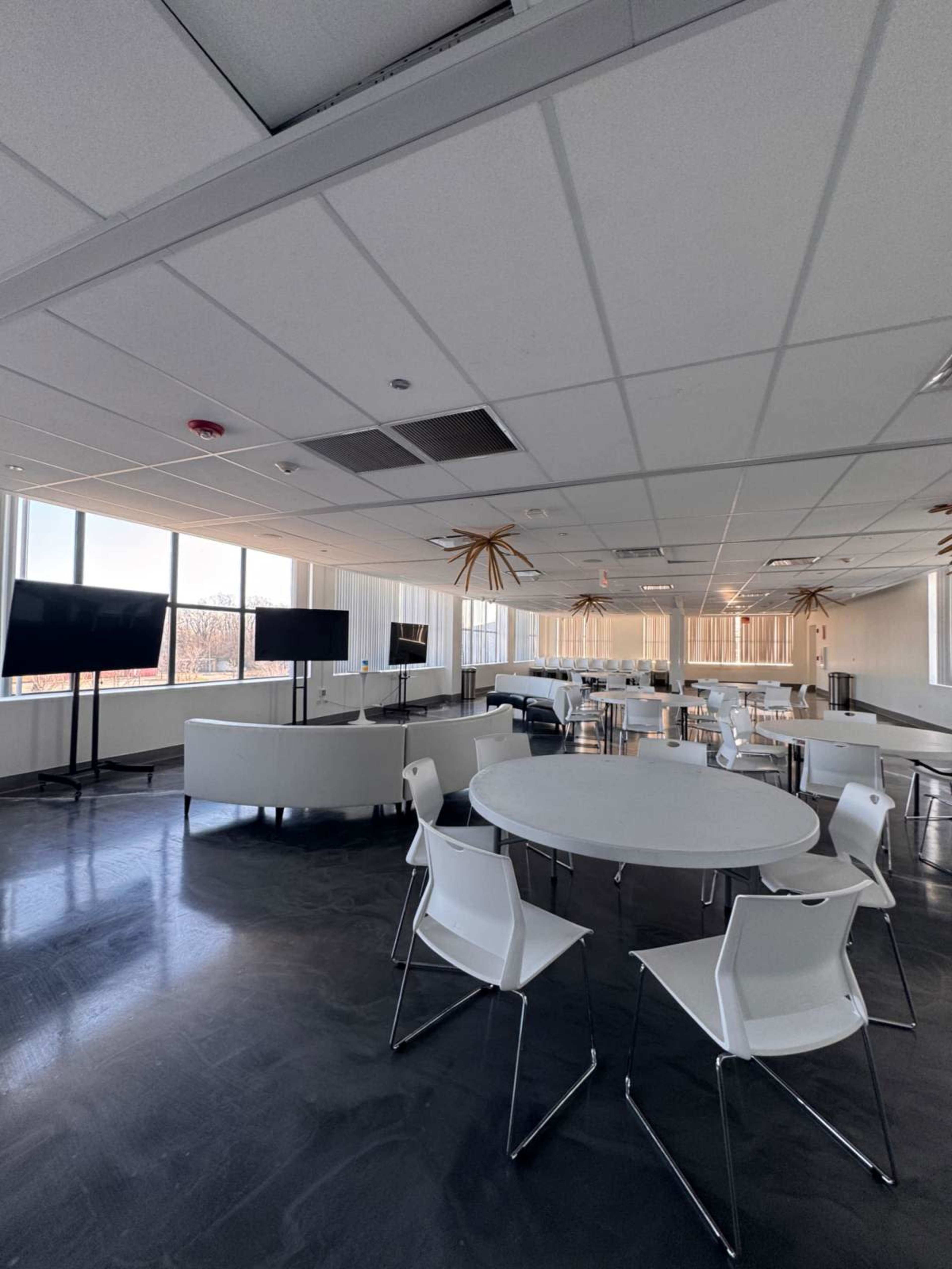 The image shows a modern, empty office space with several round tables and chairs, large windows, and two television screens mounted on stands.